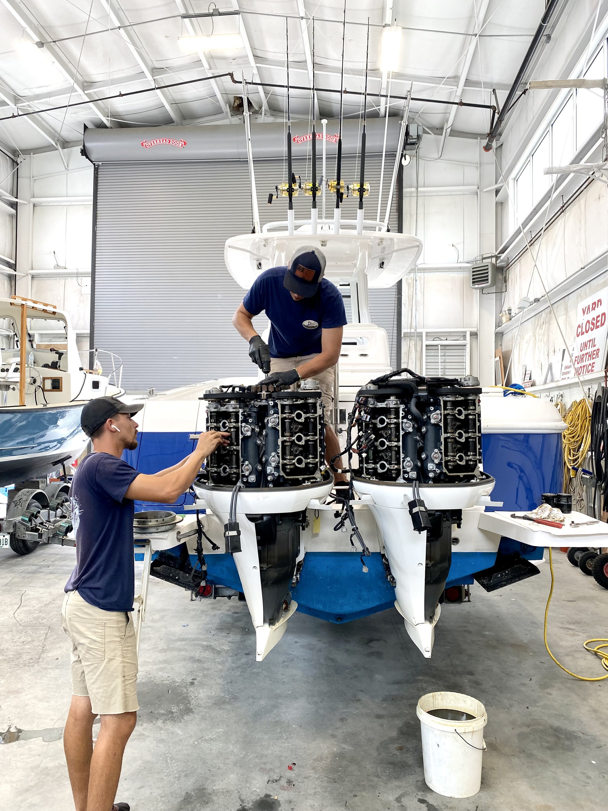 Two men working on a boat engine in a workshop, with one standing and working on the engine and another sitting nearby observing.