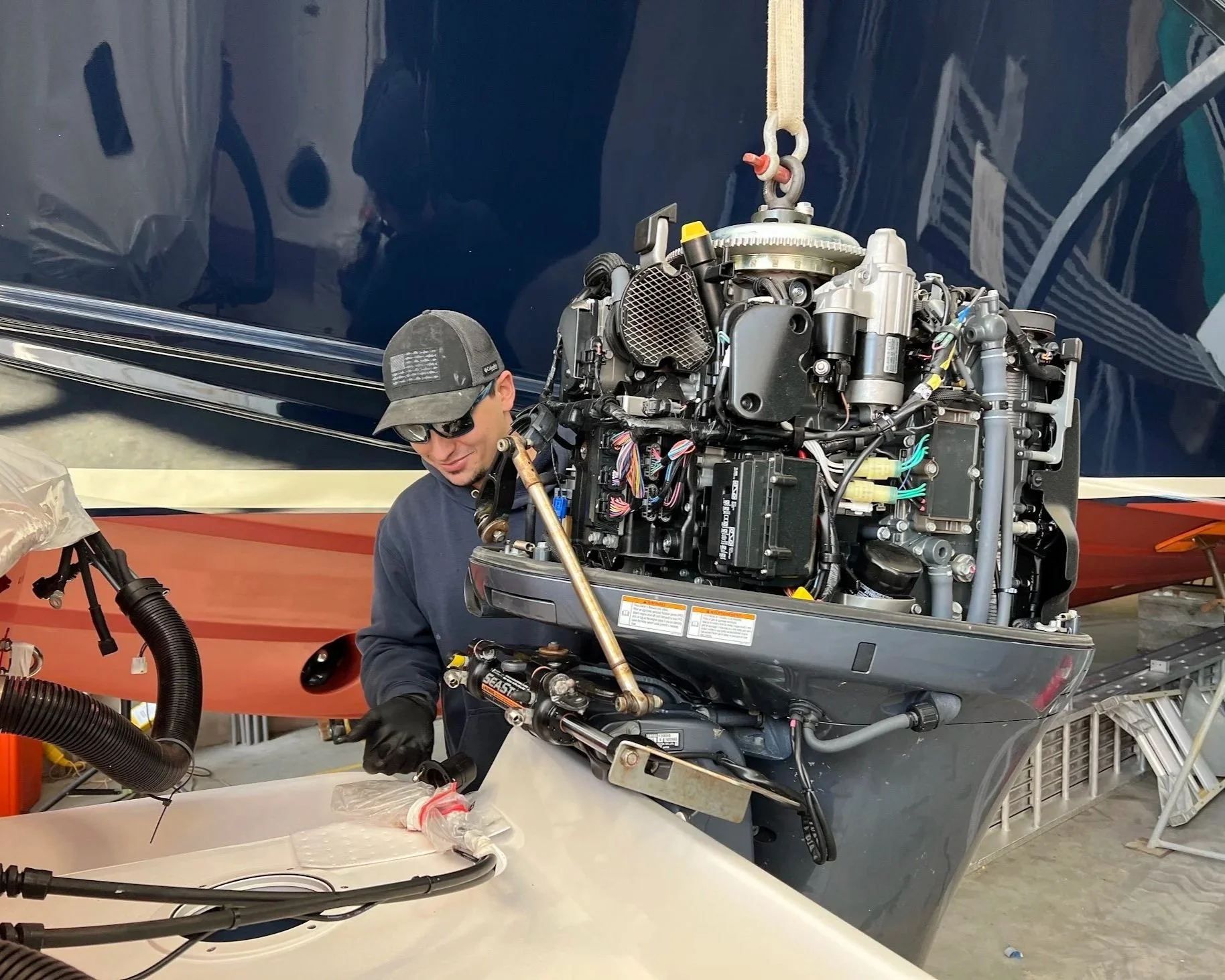 A man working on boat engine in a workshop, wearing a cap and sunglasses, with boat spray painted blue and orange in the background.