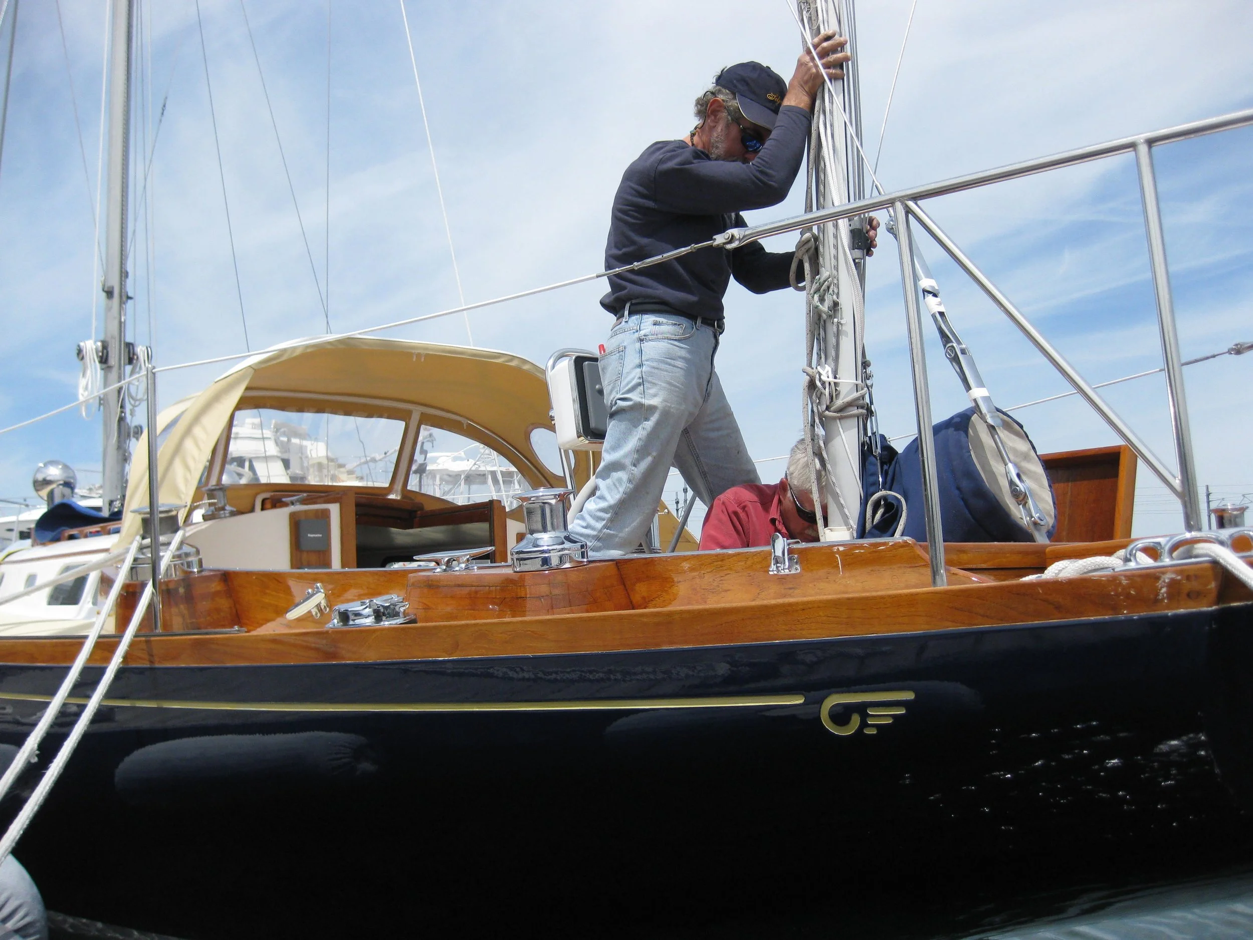 A man wearing a black cap, sunglasses, a black long-sleeve shirt, and light blue jeans is on a boat, adjusting the sails or rigging, with a woman sitting below in a red shirt. The boat has a polished wooden surface and metal railings, with a yellow canopy in the background and a clear blue sky.