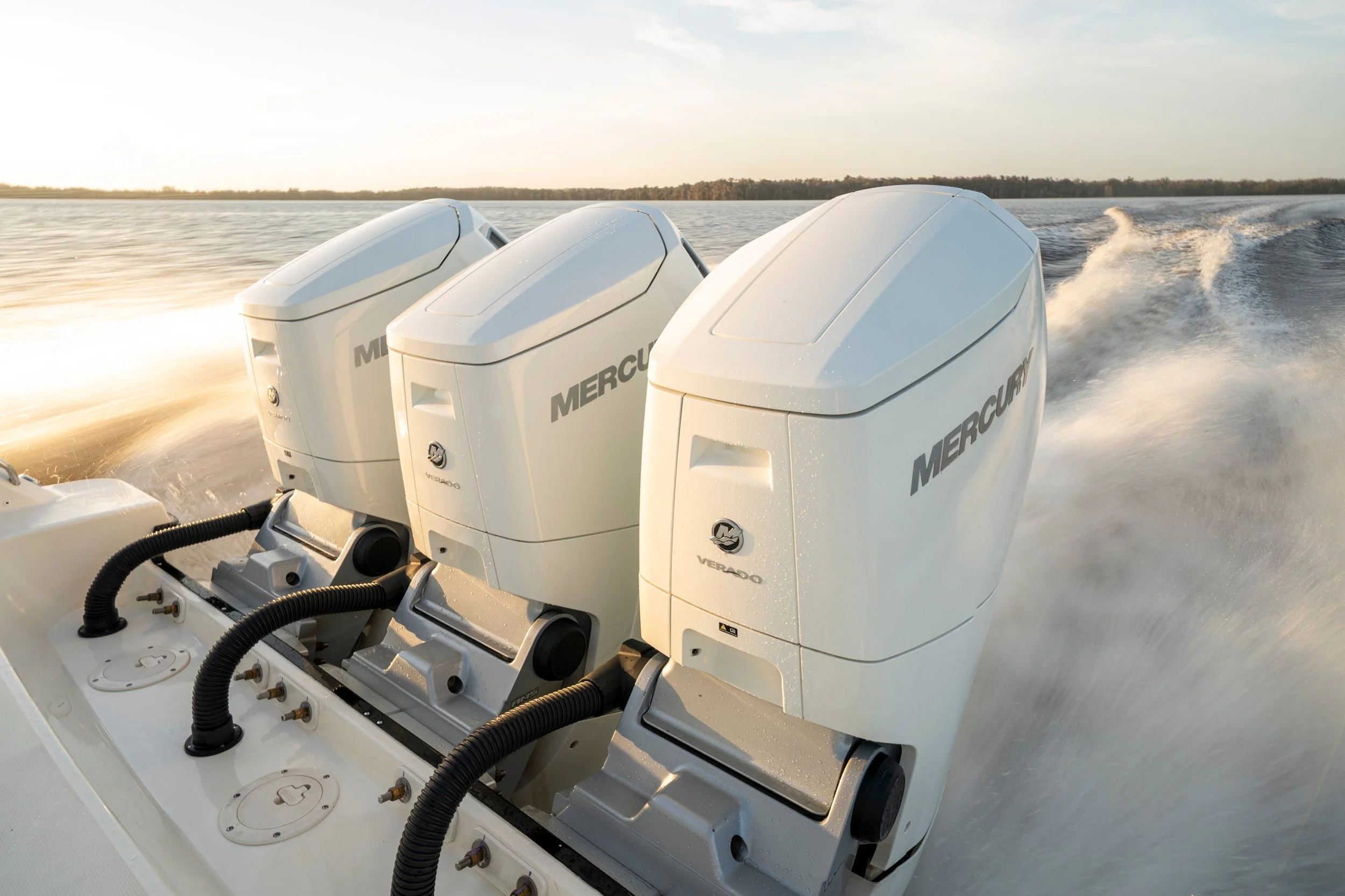 Three white Mercury outboard motors on the back of a boat moving on the water, creating a wake behind it, with a distant shoreline and a partly cloudy sky