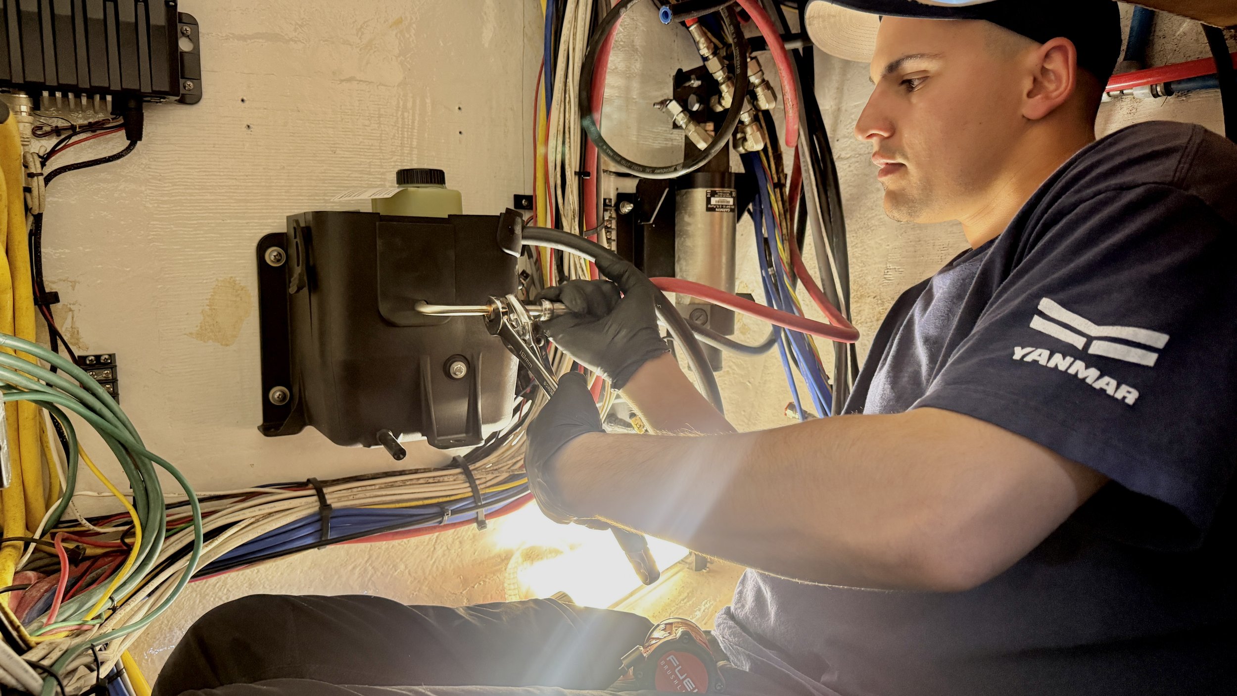 A technician working on electrical components with cables and wires around, wearing gloves and a shirt with the Yanmar logo.