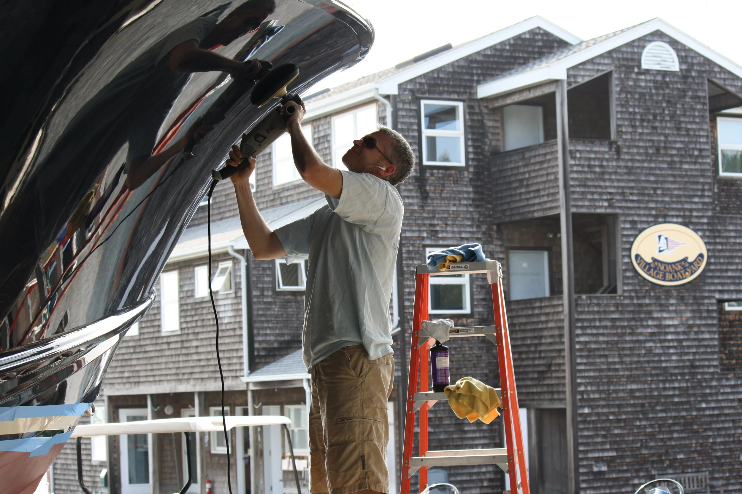 Man working on a boat, sanding or polishing its shiny surface, while standing on a ladder in front of a residential building.
