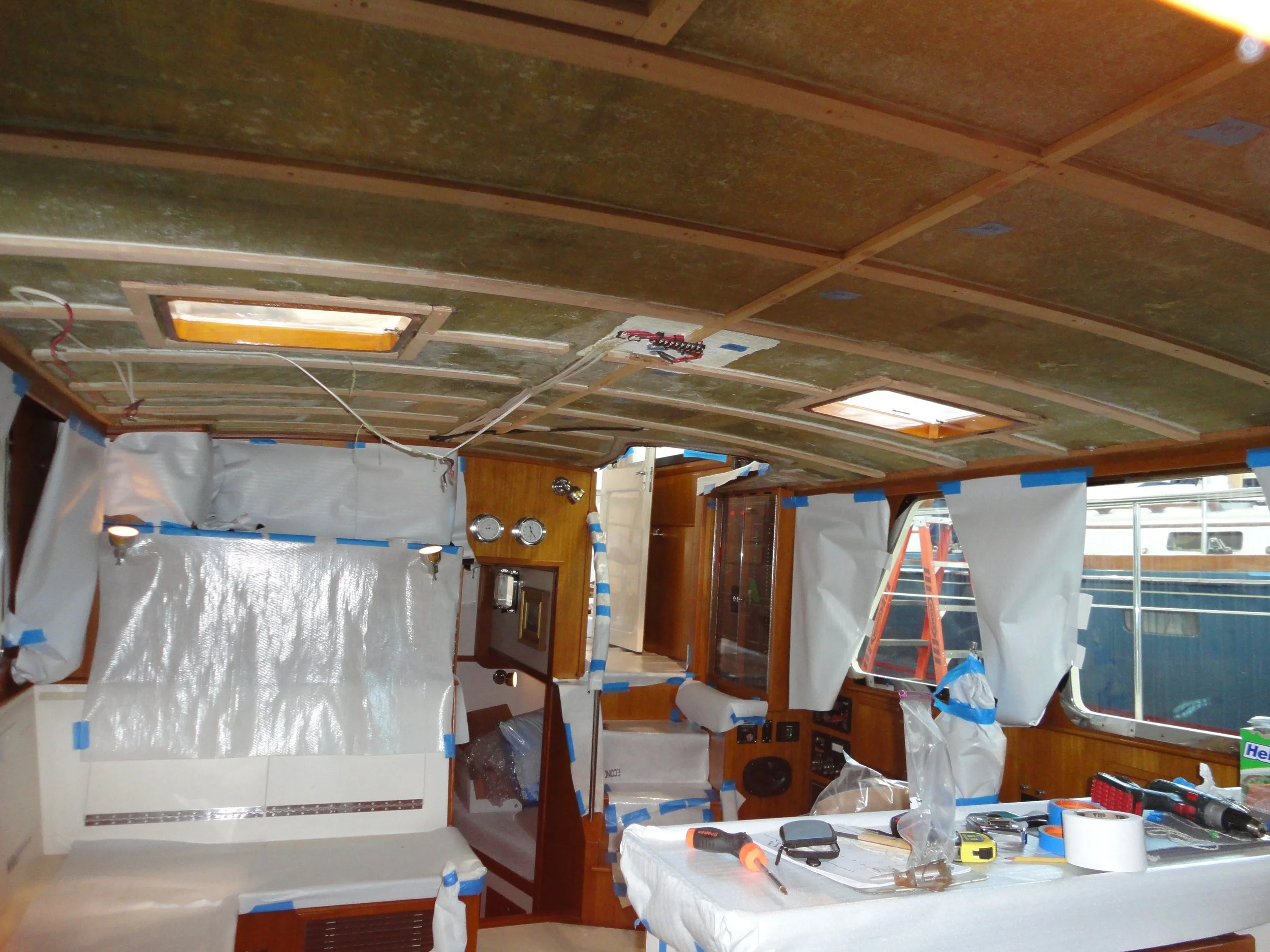 A room under construction with various tools, electrical wiring, insulation, and plastic sheeting covering surfaces. There are ceiling panels installed, and work is in progress.