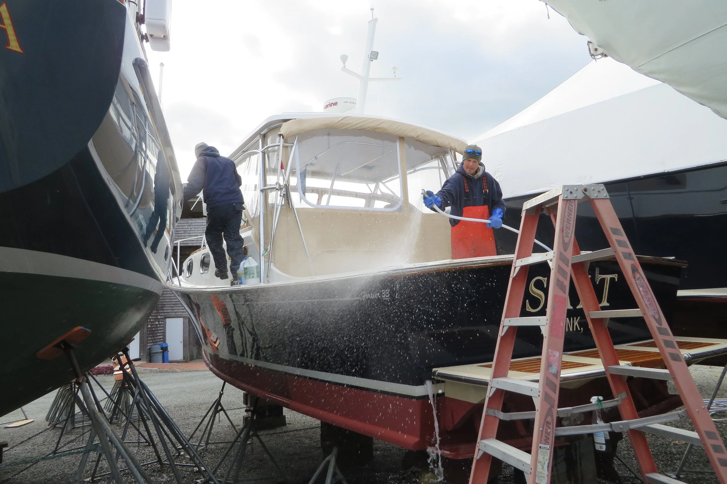Two men are cleaning a boat with a water hose while standing on a ladder among boats docked at a marina.