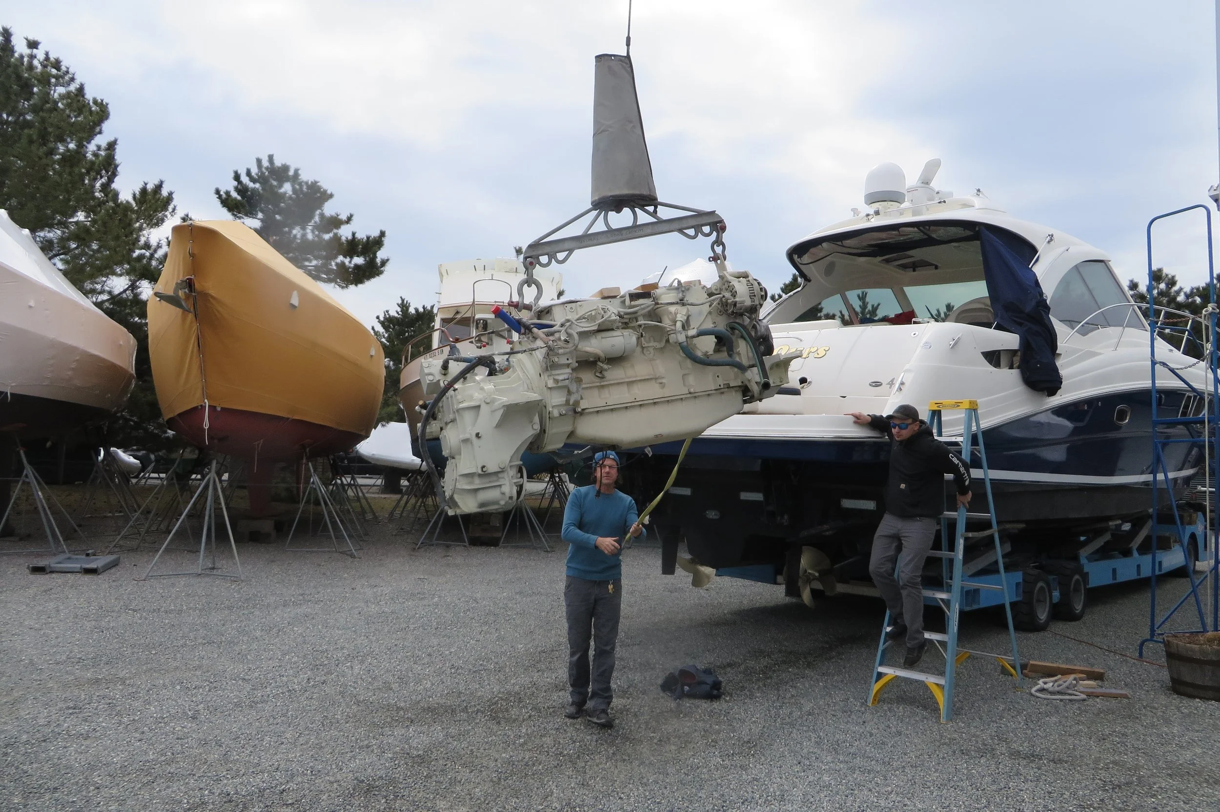 People working on maintenance or repair of boats and yacht engines at a boatyard, with boats on stands and tools around.