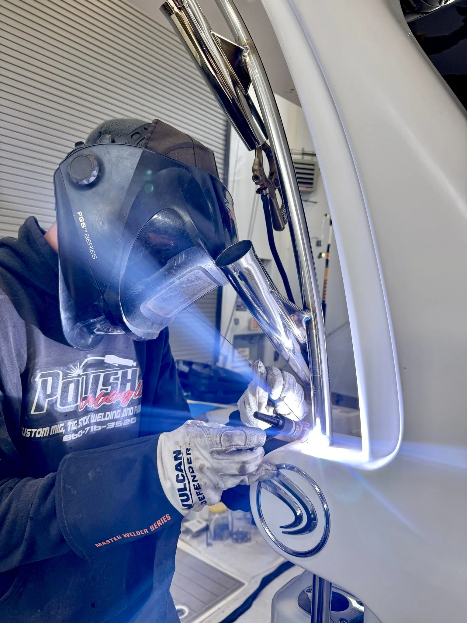 A person welding metal with a welding torch, wearing a dark shirt and welding helmet, inside a workshop.
