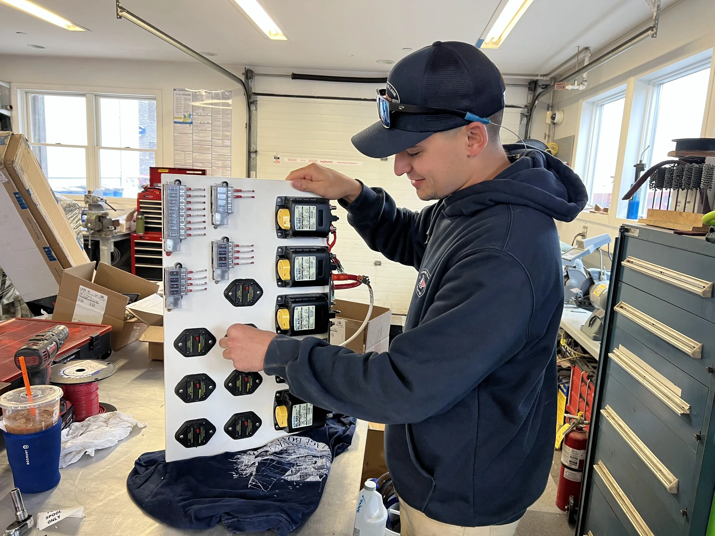 A young man in a baseball cap and sunglasses is working on an electrical control panel inside a workshop. The room has tools, boxes, and equipment on the workbenches and shelves, with large windows letting in natural light.