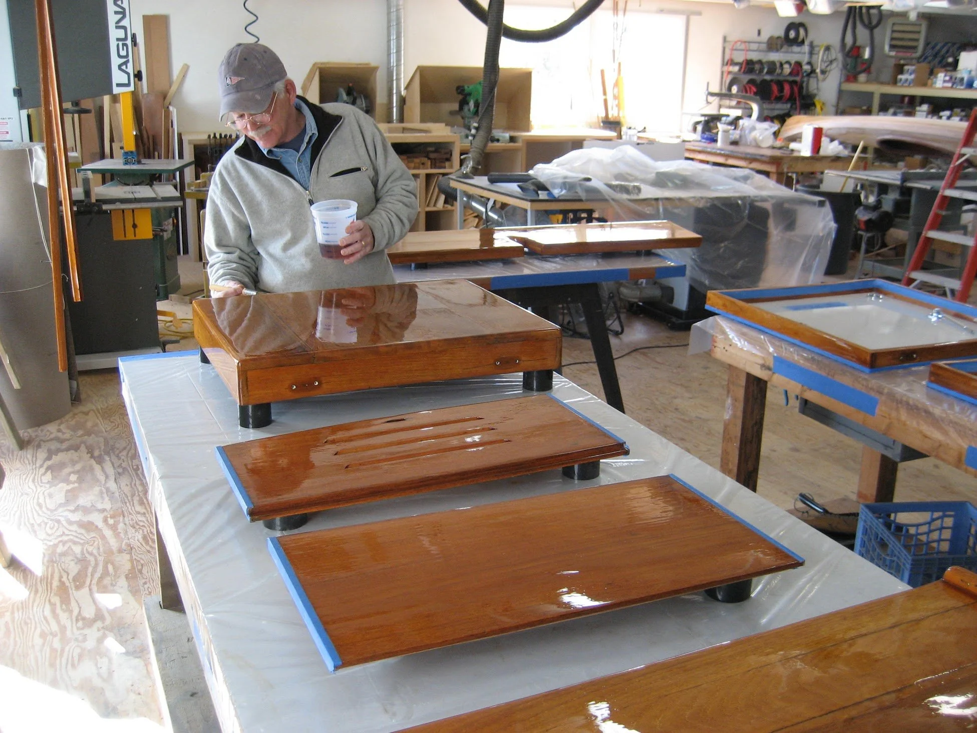 An elderly man with glasses, a gray cap, and a gray zip-up hoodie spray-tanning wooden furniture in a woodworking workshop. The workshop has various tools, racks, and shelves filled with supplies, and bright sunlight is coming through the windows.