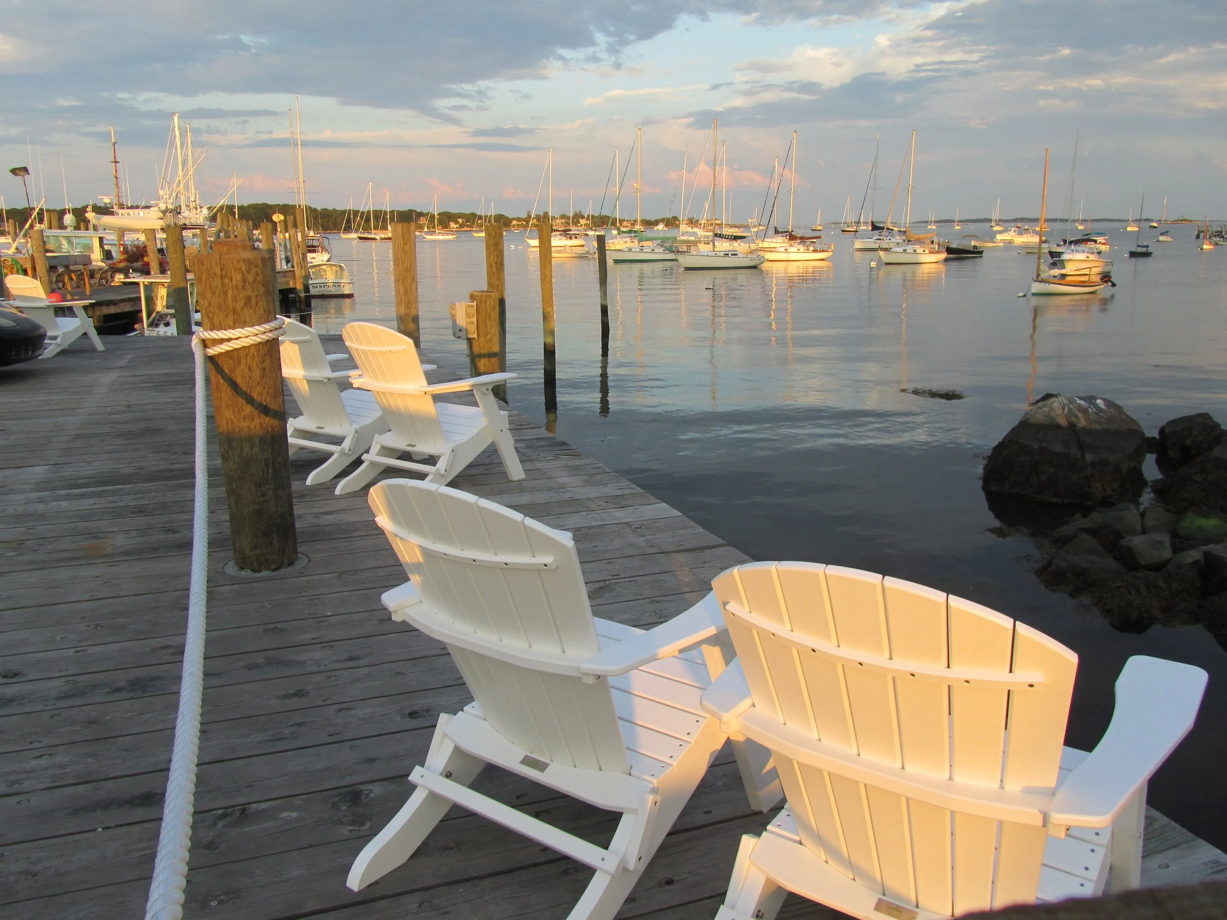 A dock with white chairs, wooden posts, and a view of boats anchored on calm water during sunset or sunrise.