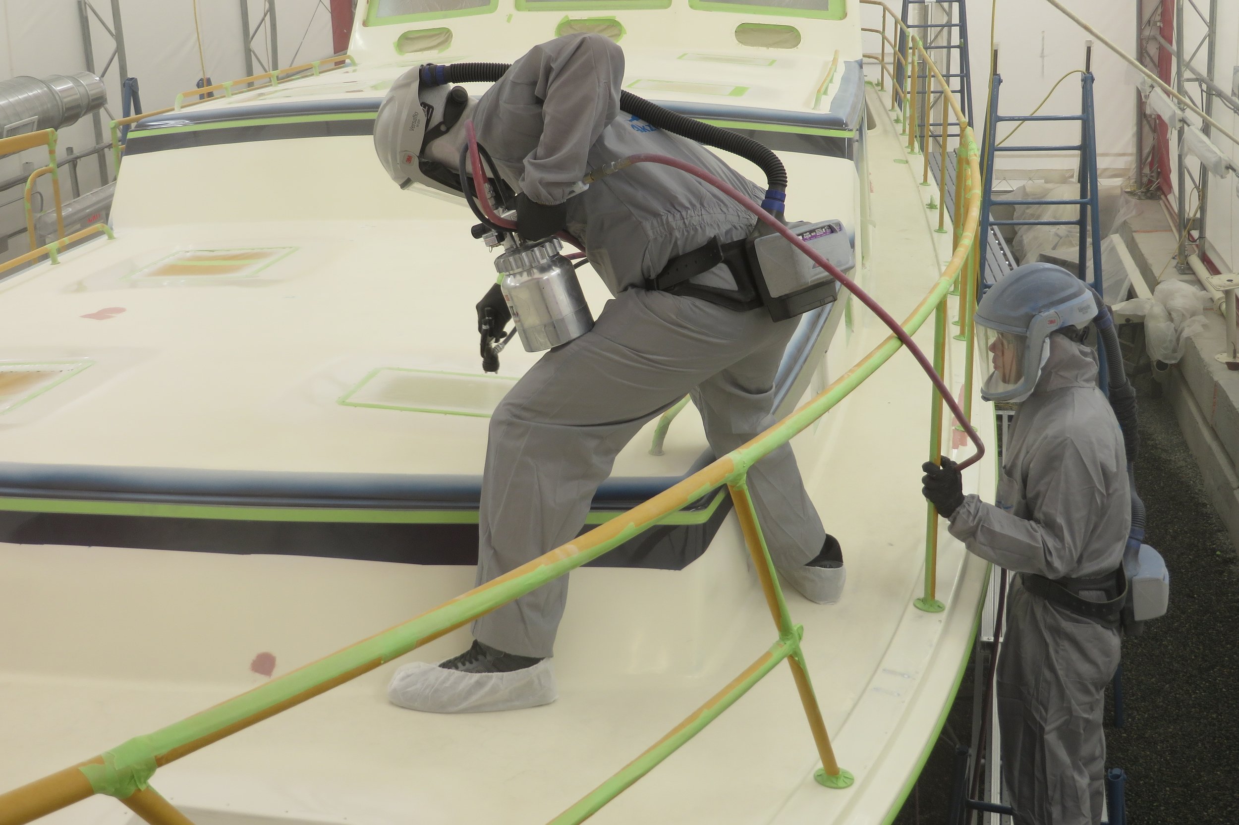 Two workers in gray protective suits, helmets, and masks are working on the exterior of a spacecraft or aircraft, applying paint or finish work on its surface inside a warehouse or hangar.