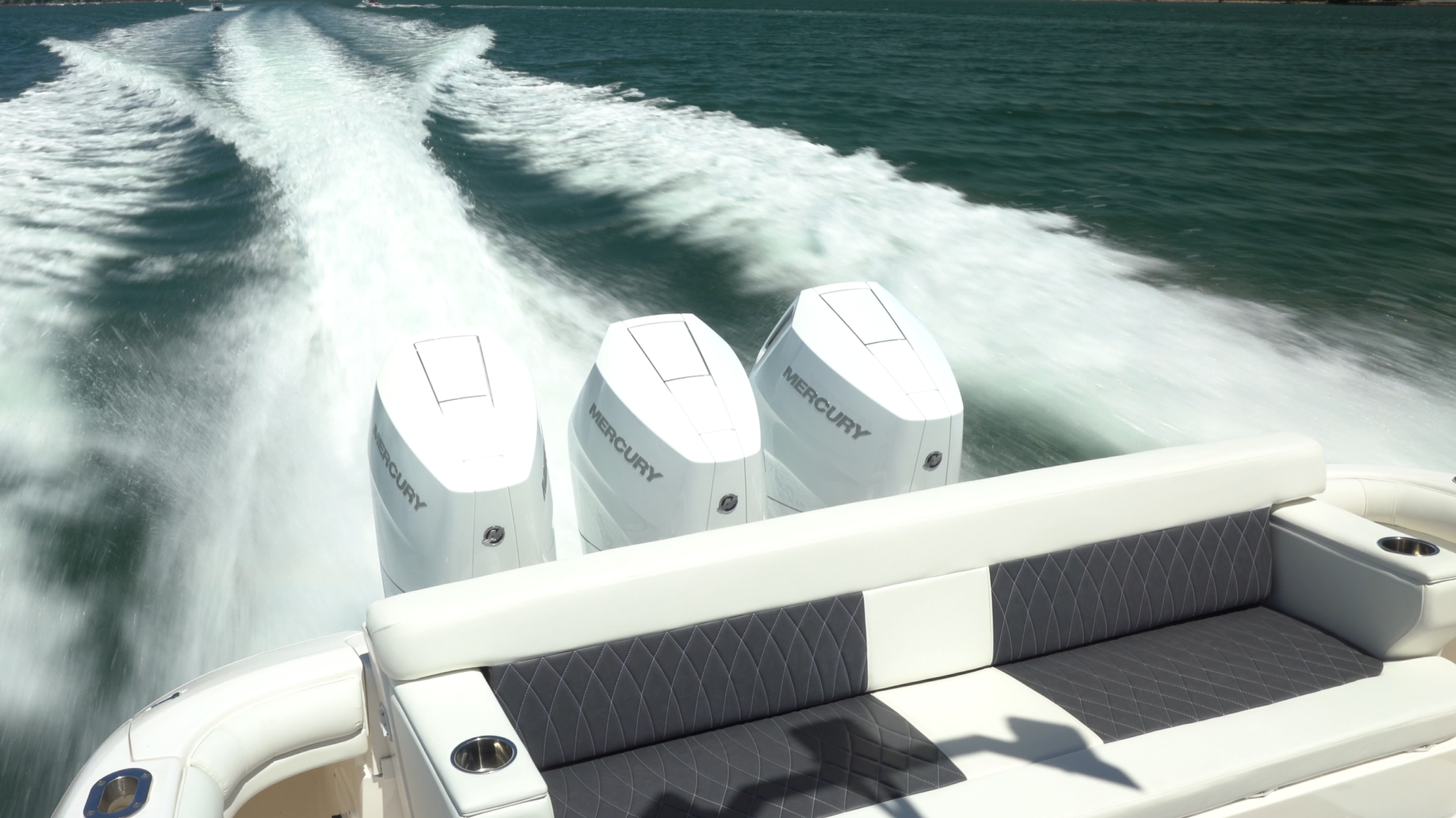 Back of a motorboat with four Mercury outboard engines, moving through water and creating a wake.