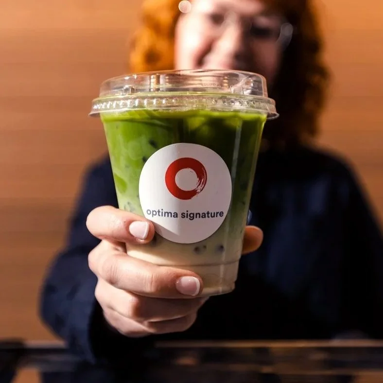 A woman holding a plastic cup of green and white bubble tea with a clear plastic lid and a sticker that says "optima signature." The woman is smiling with a blurred wooden wall background.