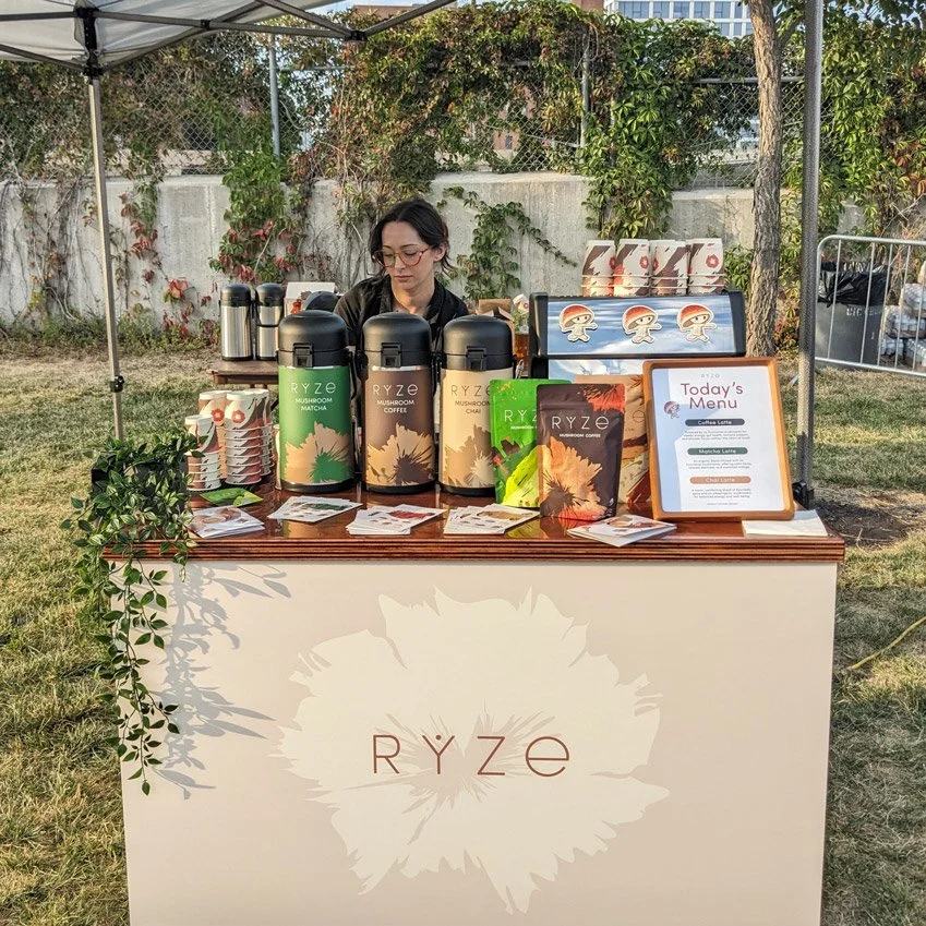 Front view of the cart branded for Ryze Coffee, with coffee pots on the cart table and a woman standing behind outside on grass