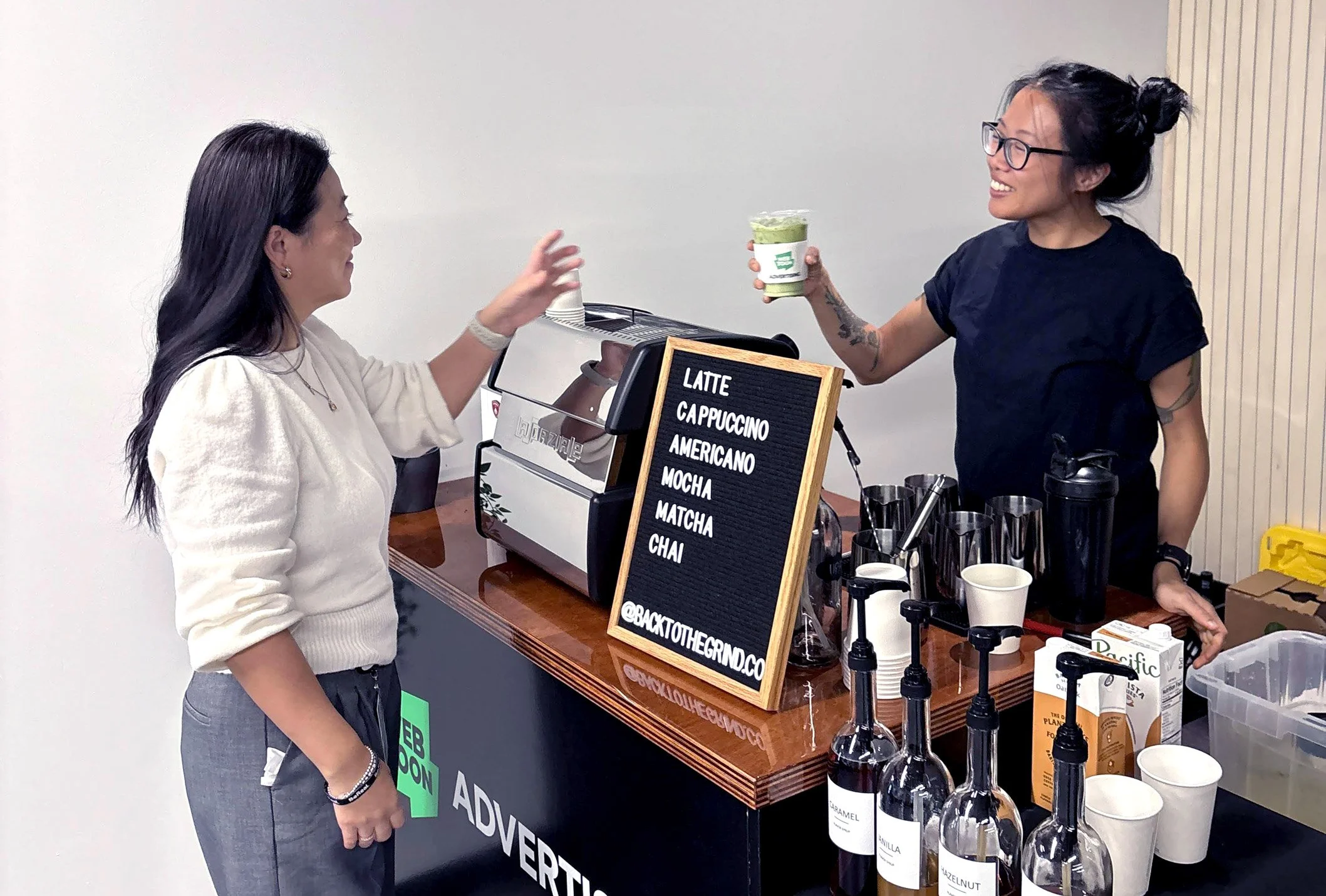 Two women at a coffee stand with a black menu board listing coffee types, one woman handing the other a green beverage in a cup.