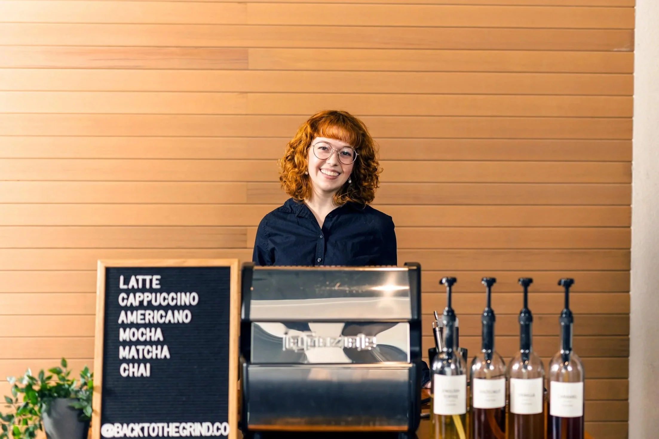 A smiling woman with red hair, wearing glasses and a dark blue shirt, standing behind a coffee stand with a menu sign listing popular drinks like latte, cappuccino, and mocha, in a setting with a wooden wall background and bottles of beverage on the counter.