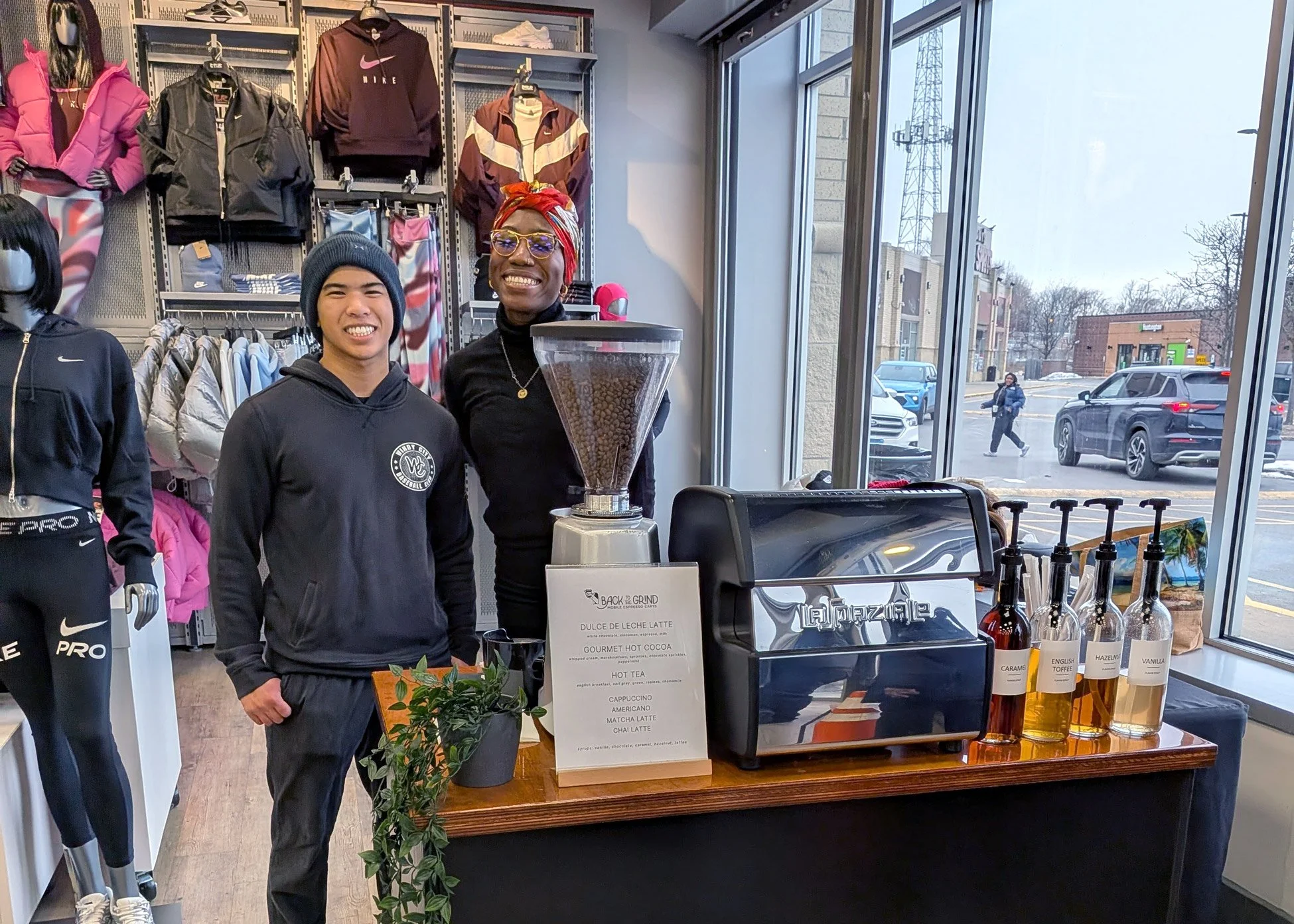Two people behind the coffee cart set up in a clothing store smiling for the camera