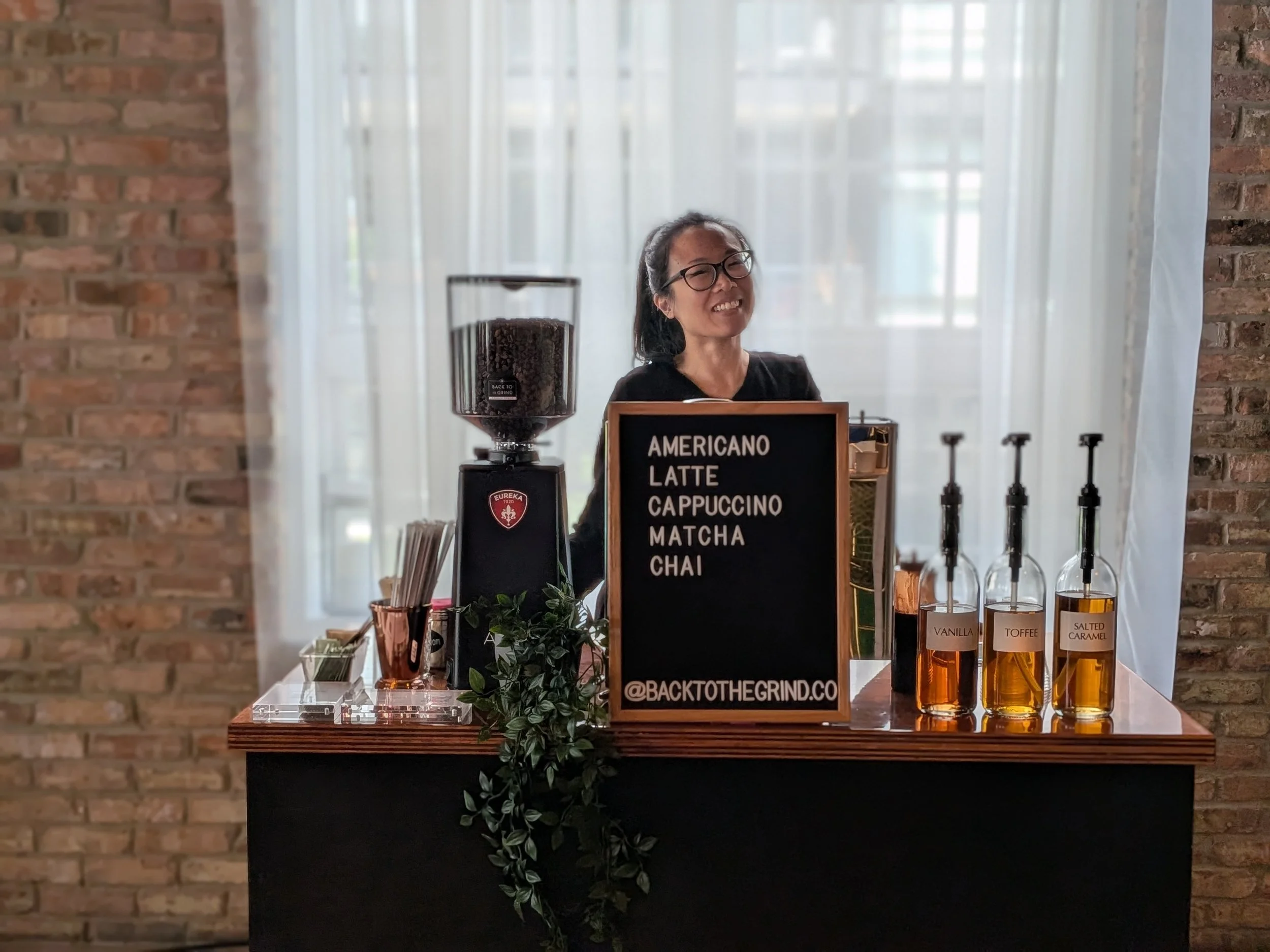 A woman smiling behind a coffee stand menu with a variety of coffee drinks listed, including Americano, Latte, Cappuccino, Matcha, and Chai. The stand features a large coffee grinder, bottles with flavor syrups, and a decorative green plant, all set against a background of white curtains and brick walls.