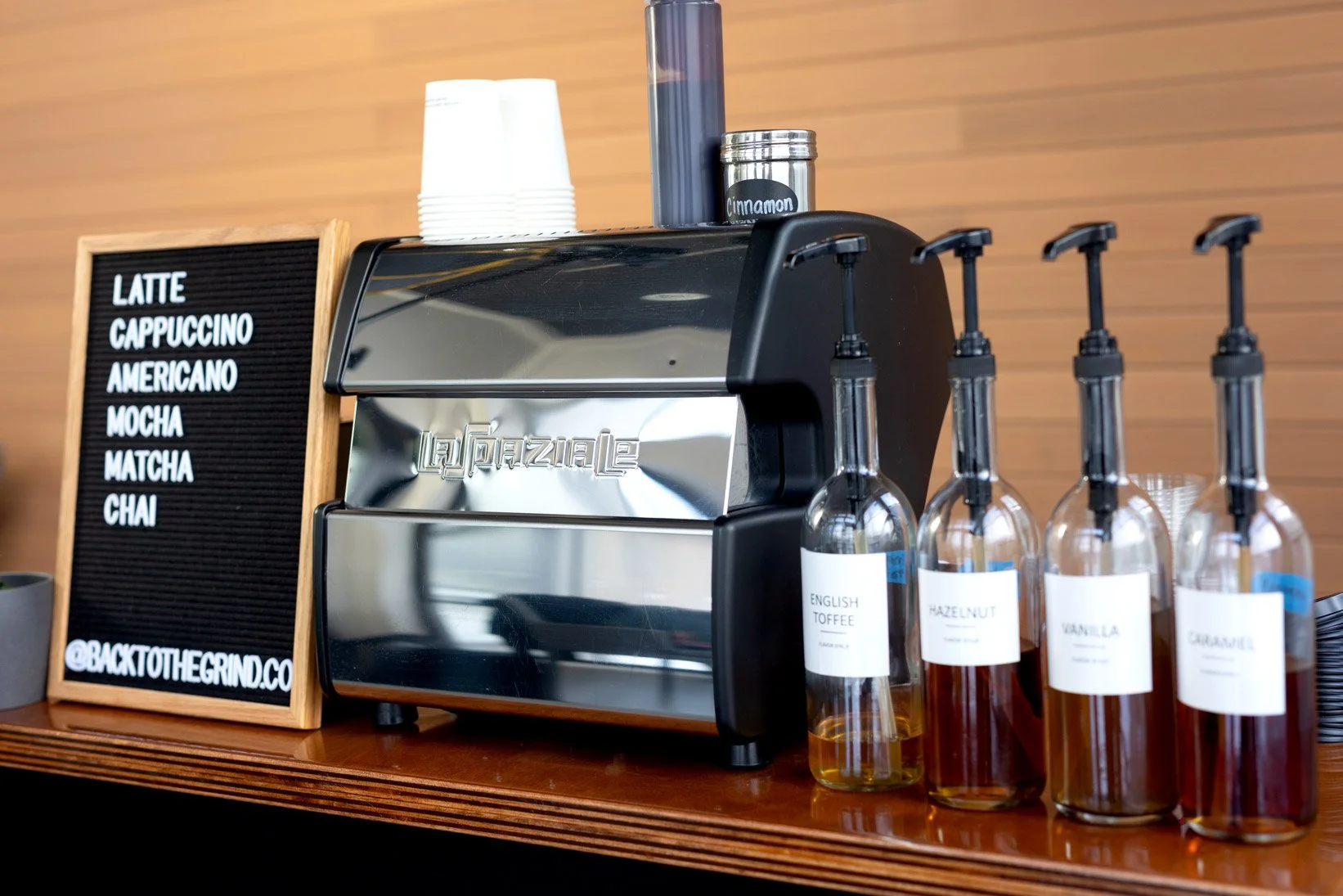 A coffee counter with an espresso machine, a list of coffee drinks on a blackboard, and bottles of flavored syrup labeled English Toffee, Hazelnut, Vanilla, and Caramel. The background has a wooden wall.