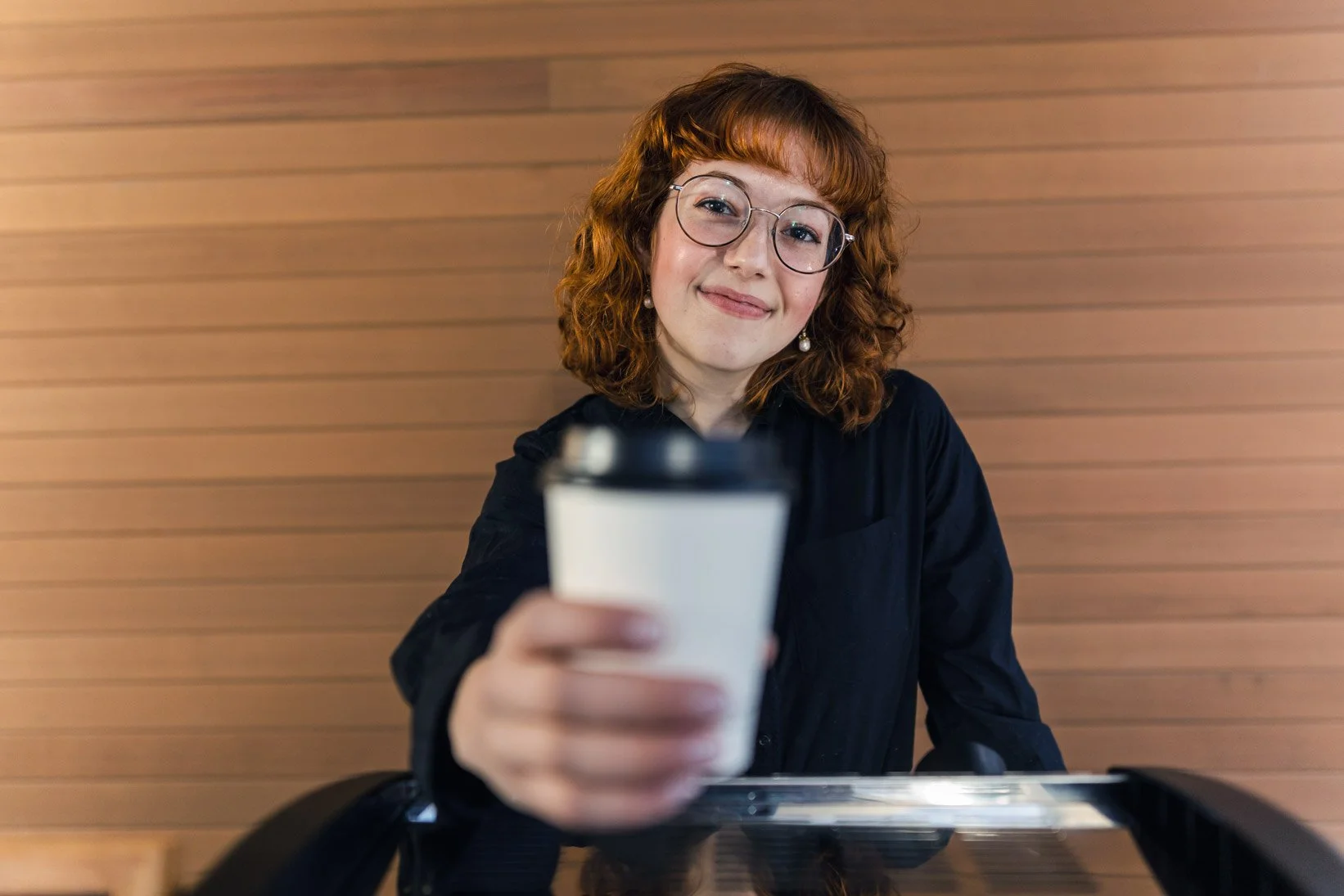 A female barista with red curly hair, glasses, and earrings smiling while holding a coffee cup towards the camera, sitting at a table against a wooden wall.