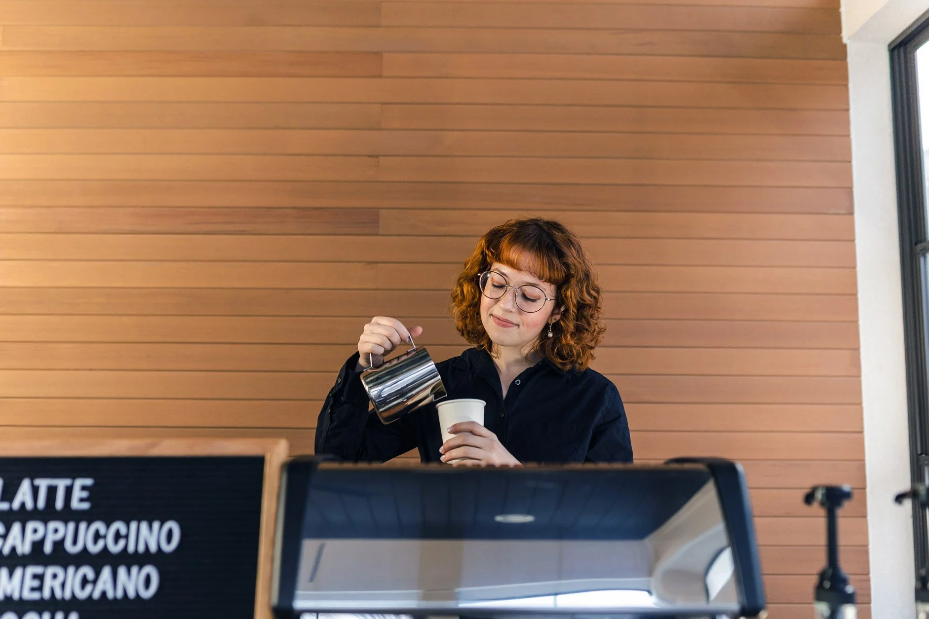A woman with curly brown hair and glasses pouring coffee from a metallic pot into a paper cup at a cafe with a wooden wall background.