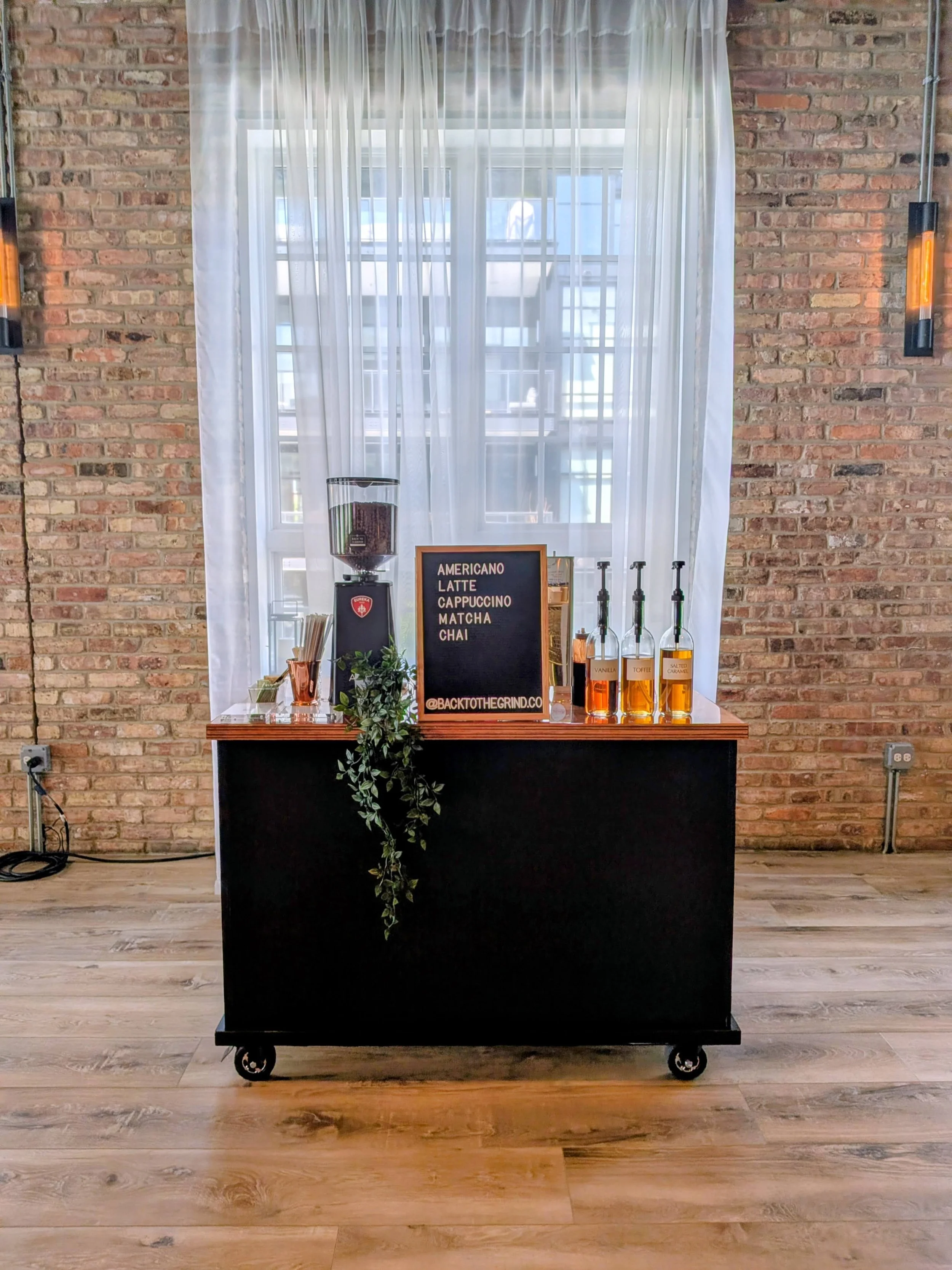A black mobile bar cart on wheels with a wooden top, decorated with a plant, a small blackboard menu, and bottles of flavored syrup, set against a brick wall with a white curtain and a window behind it.