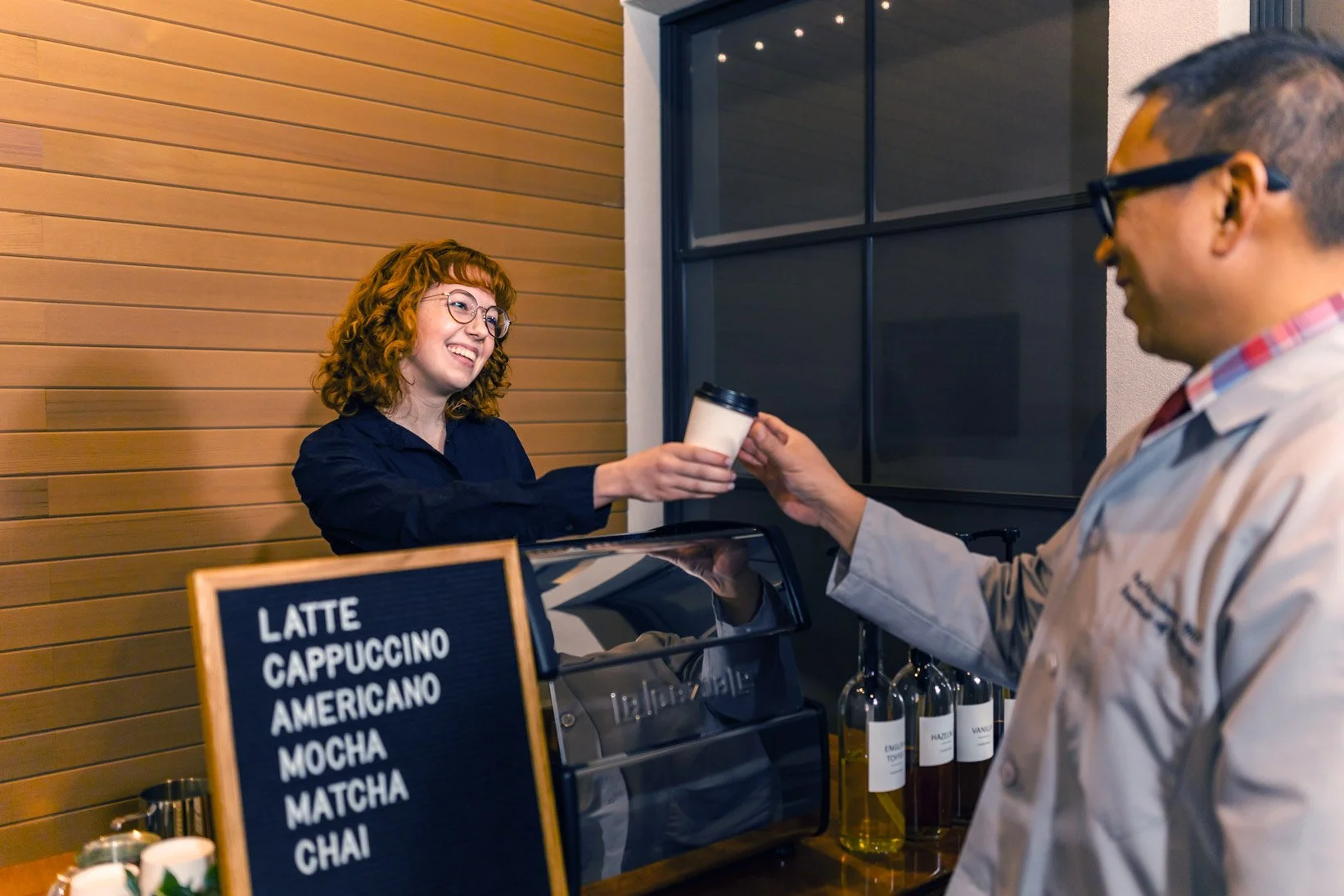 A woman in a dark shirt is smiling as she hands a cup of coffee to a man wearing glasses and a beige jacket at a coffee shop.