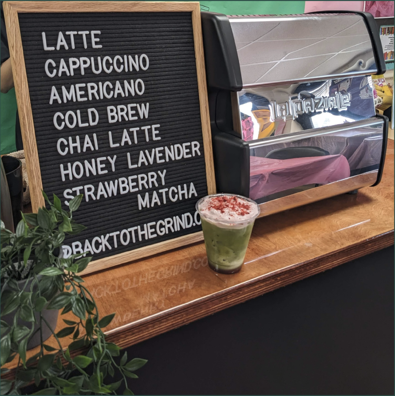 A black felt letter board with a wooden frame listing drink options: latte, cappuccino, americano, cold brew, chai latte, honey lavender, strawberry, matcha. The board is on a wooden counter next to a green plant. A clear plastic cup with a green drink topped with red powder sits in front of the board. A shiny espresso machine is behind the board.