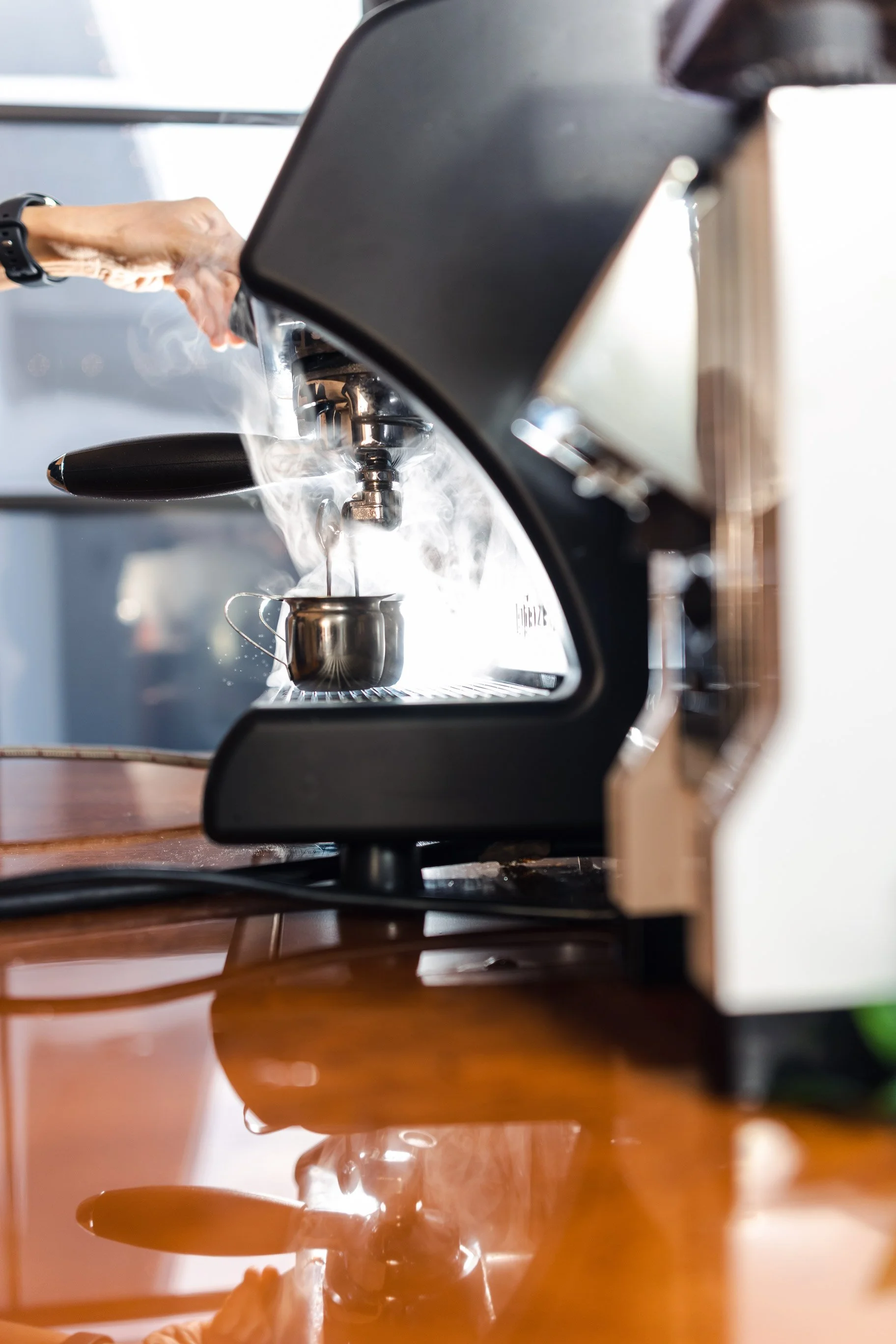 A close-up view of a commercial espresso machine with a barista hand operating it, brewing coffee into a stainless steel cup with steam rising, taken through a mirror on a wooden table surface.