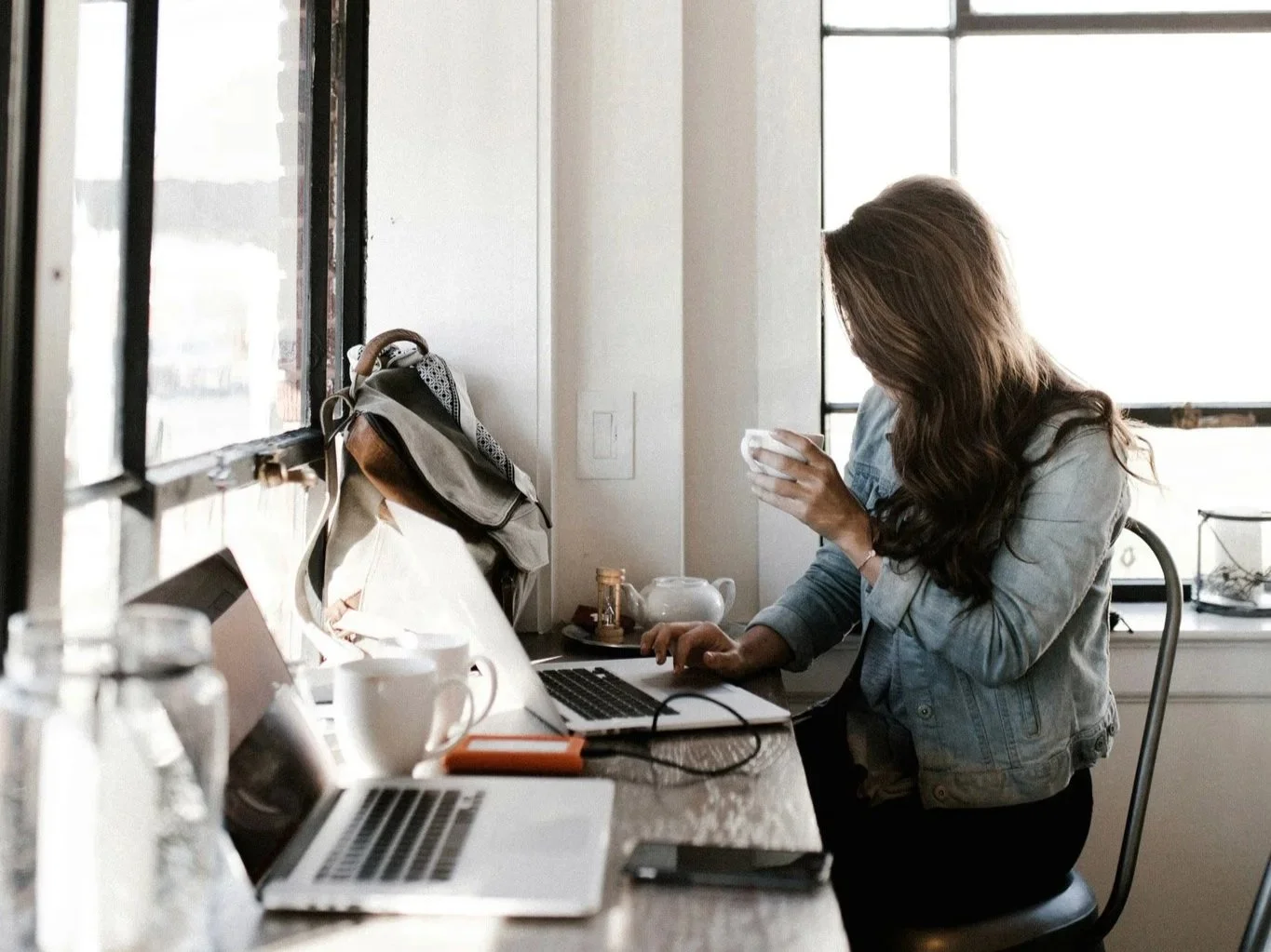 Woman with long hair in a denim jacket sitting at a cluttered desk, looking at her phone in a bright room with large windows.