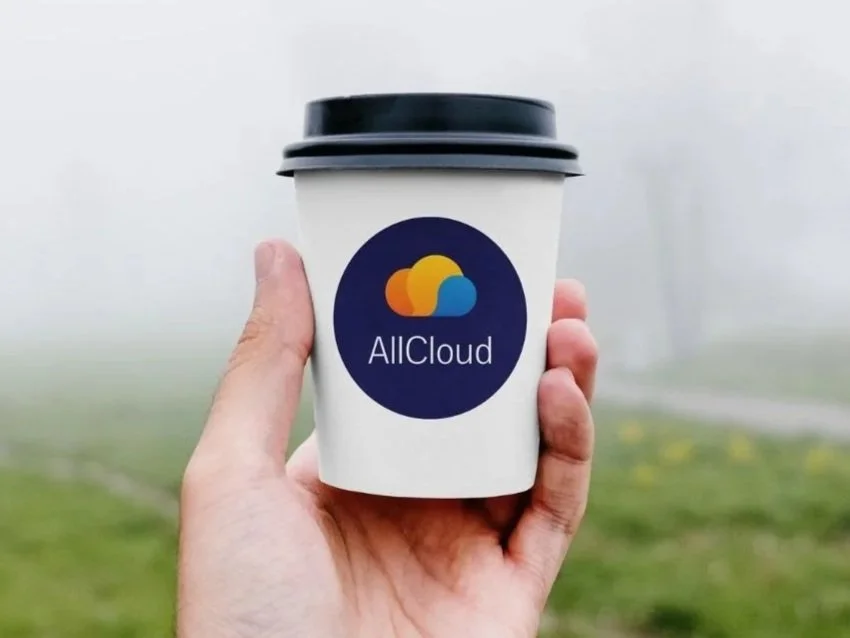 Hand holding a disposable coffee cup with an AllCloud logo, outdoors with foggy background.
