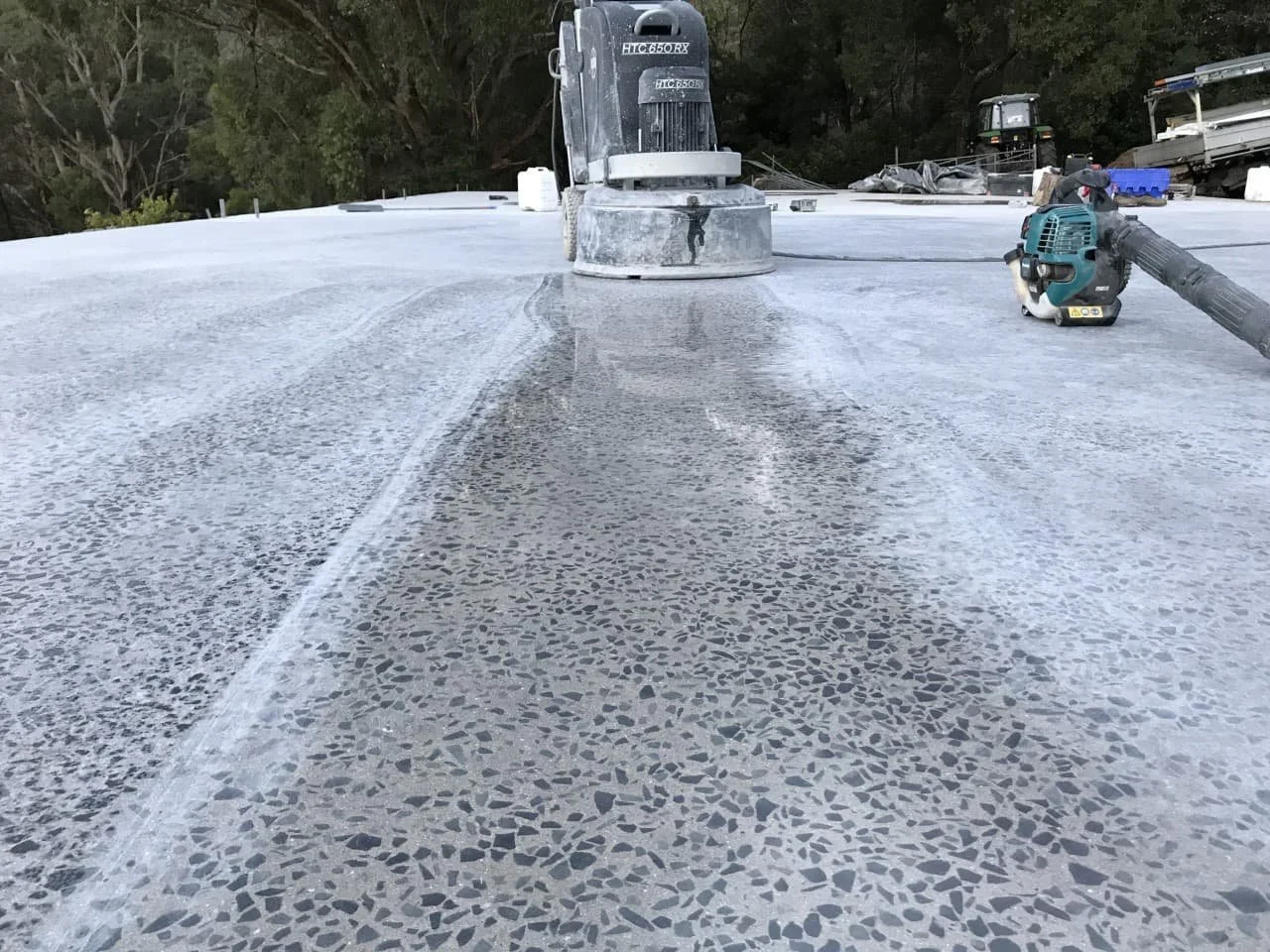 Close-up of a flat roof surface with a large concrete grinding or polishing machine, a small power tool, and other equipment, with trees and a cloudy sky in the background.