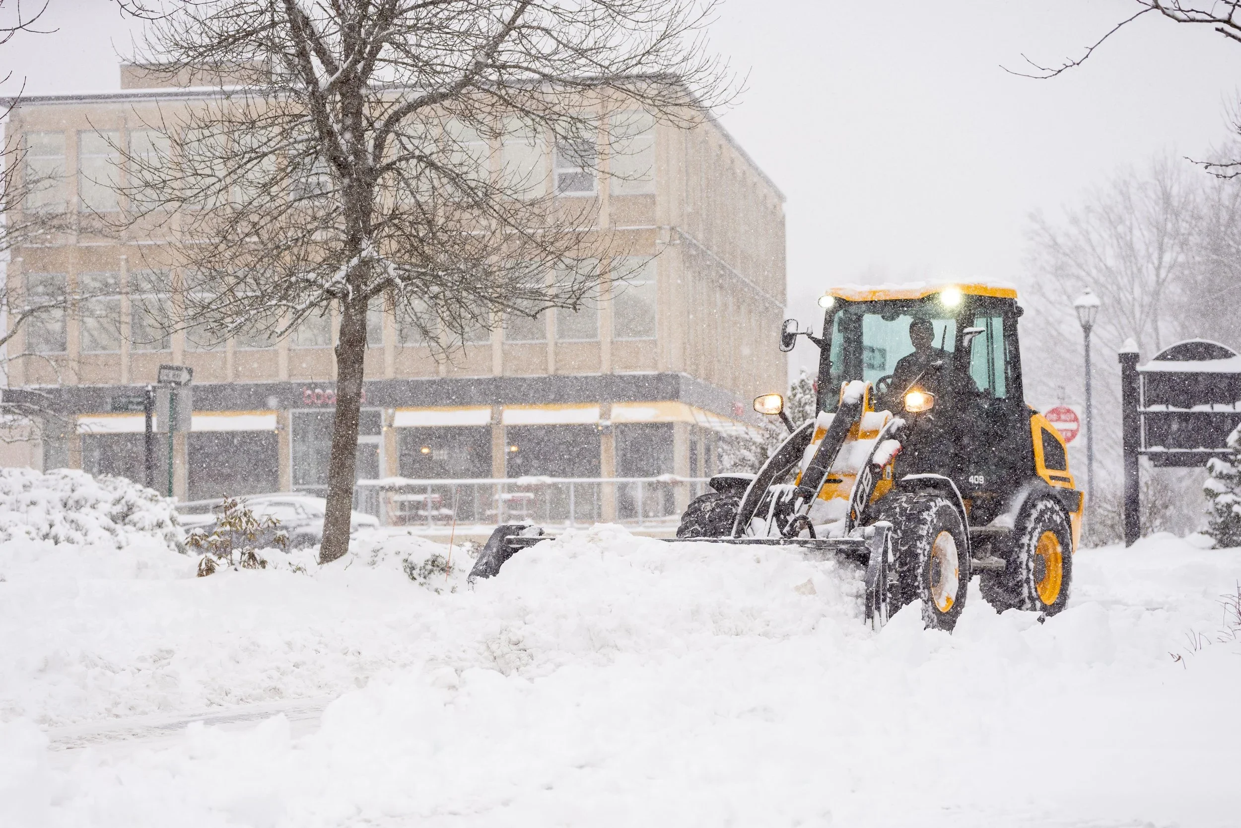 A yellow snowplow clearing snow in an urban area during a snowstorm with snow-covered trees and buildings in the background.