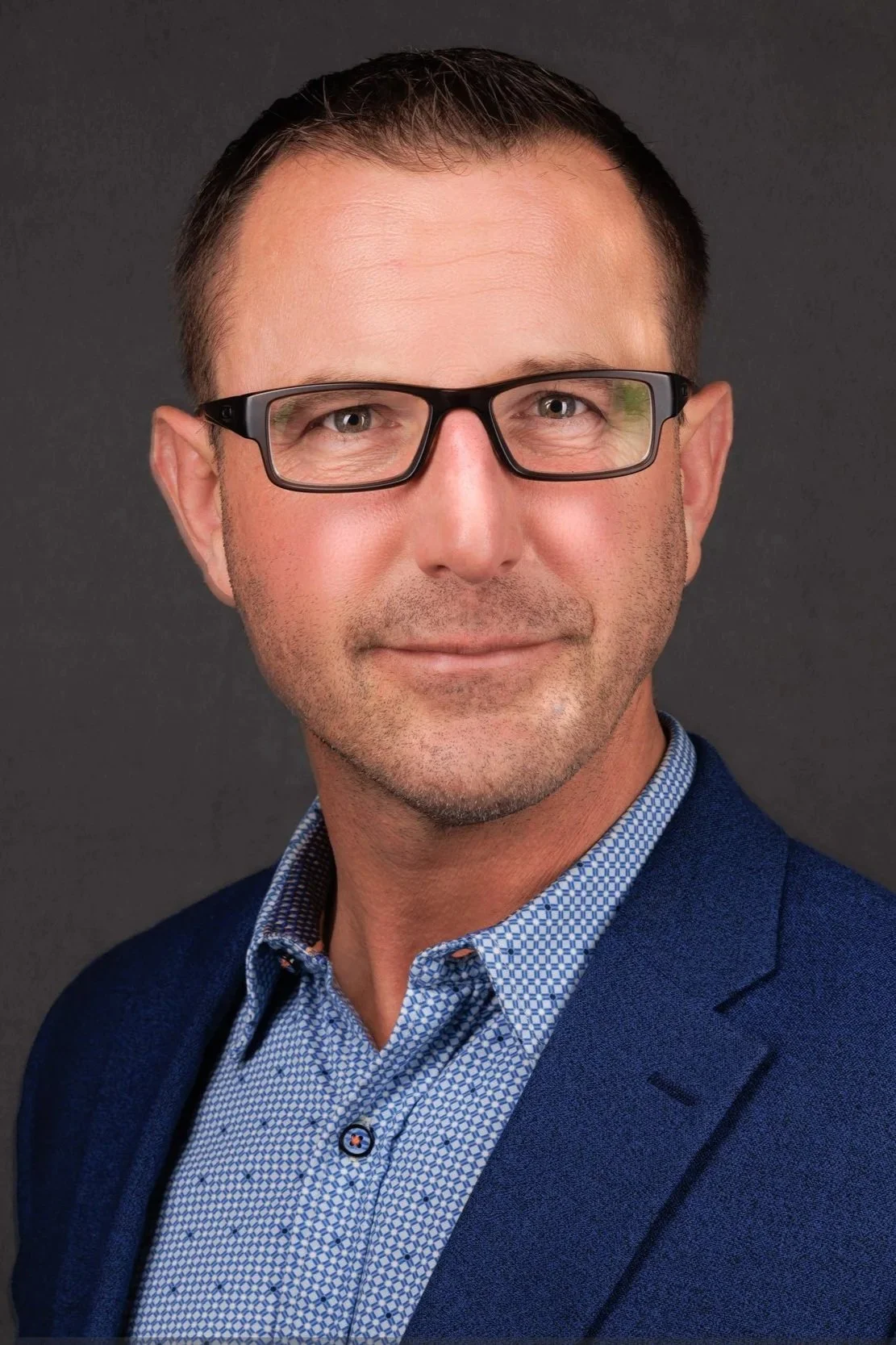 A man wearing glasses, a blue blazer, and a light blue patterned shirt, posing against a gray background.