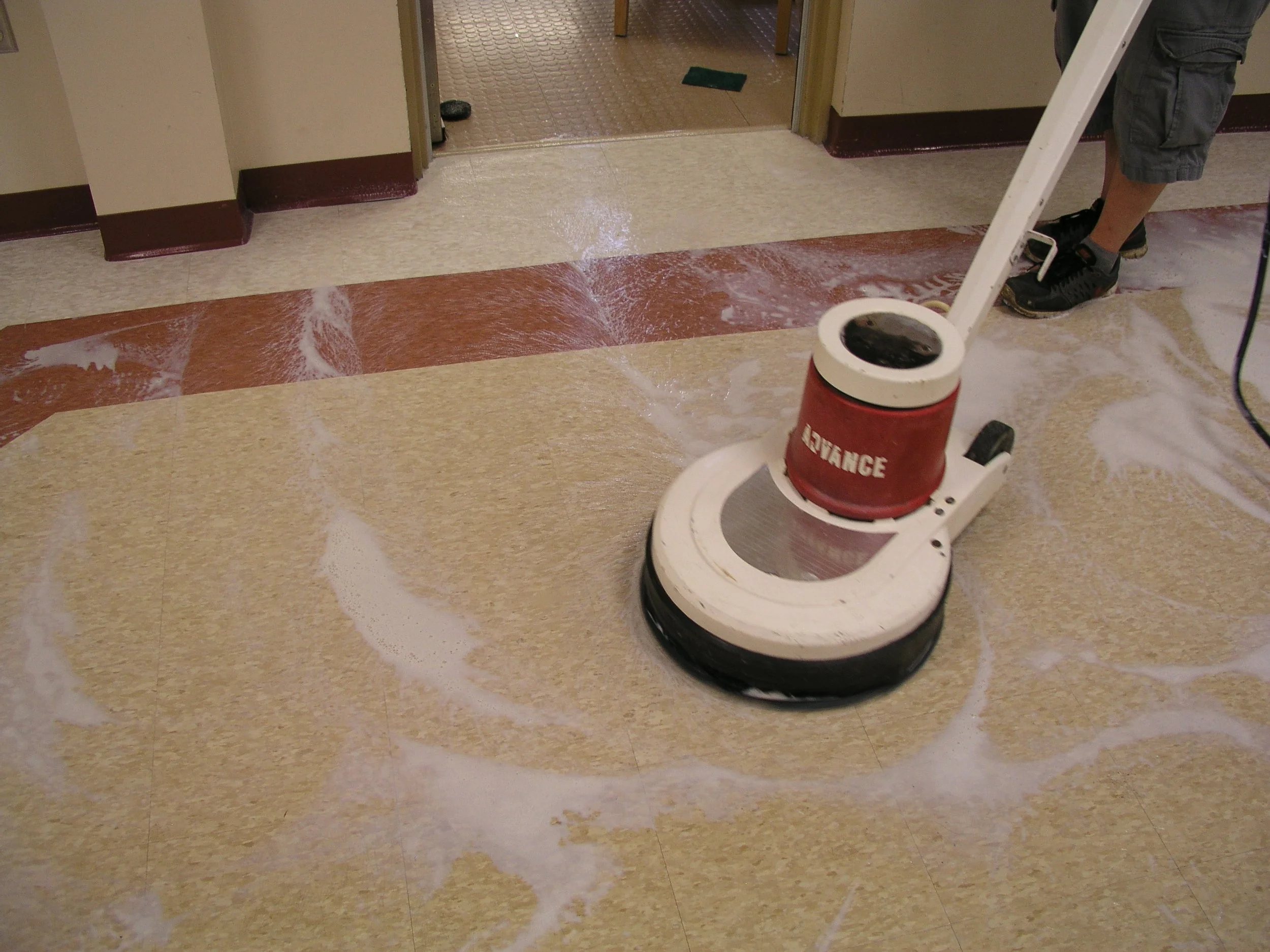 A person cleaning a tiled floor with a floor buffer machine, which has soap or cleaning solution on the floor.