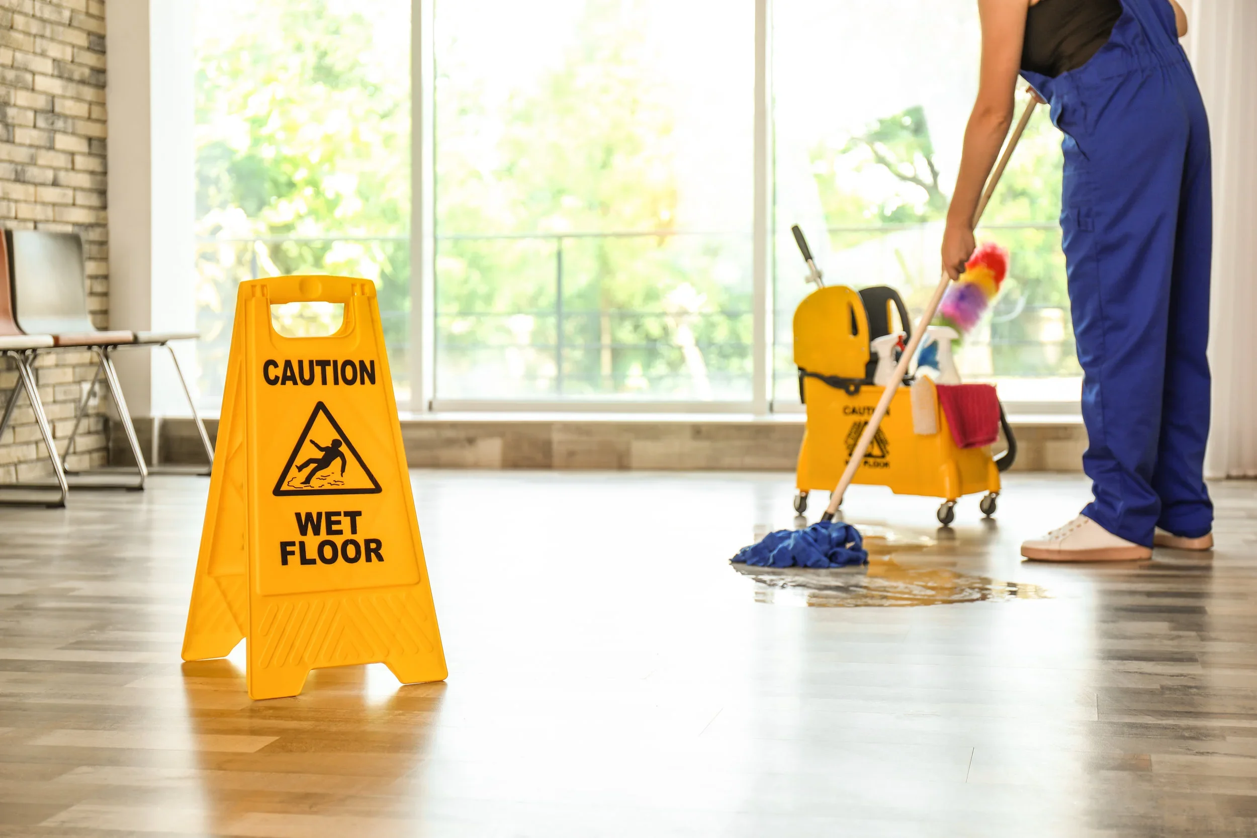 A person mopping a wet floor indoors near large windows. A bright yellow caution sign indicating a wet floor is prominently displayed in the foreground.