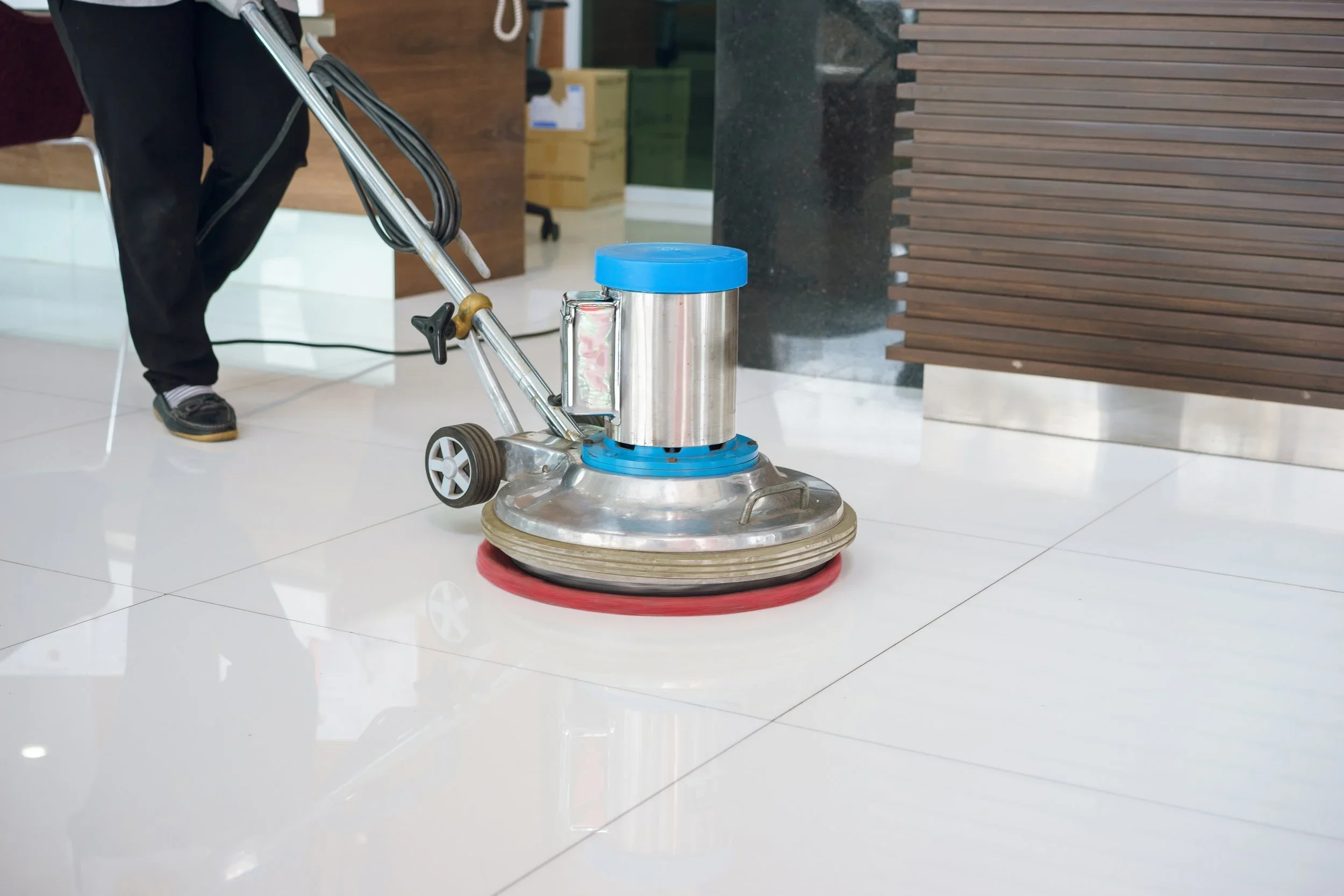 A commercial floor polishing machine in operation on a shiny tiled floor, with part of a person's leg visible in the background, situated near a wooden-paneled wall.