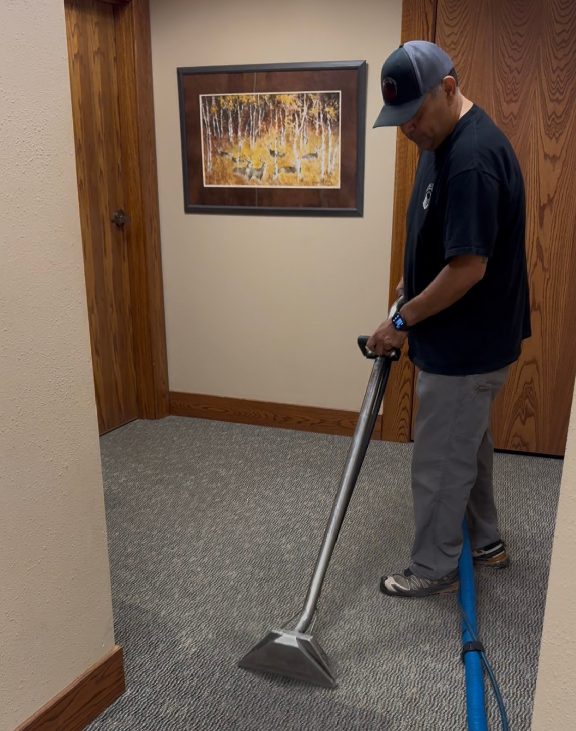 A man standing on a carpeted floor, wearing a dark T-shirt, gray pants, sneakers, and a gray cap, using a blue vacuum cleaner to clean the carpet in an office or waiting room area.