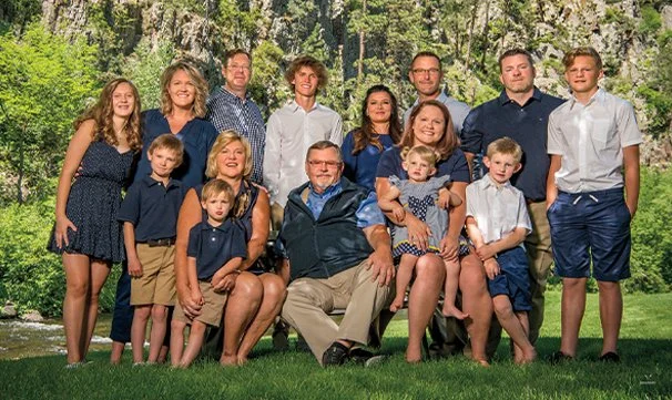Family group photo outdoors in front of trees, with adults and children dressed in casual summer clothing.