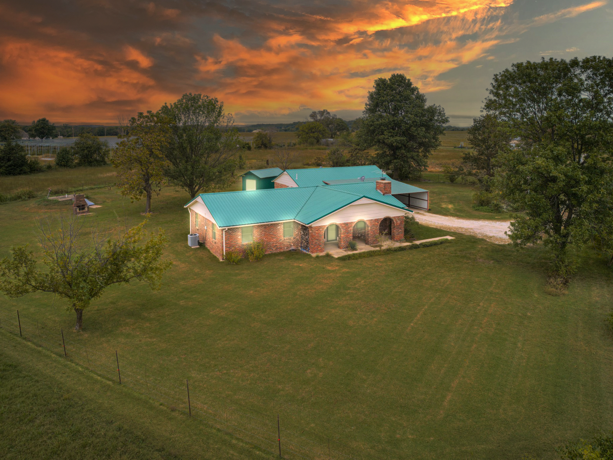 A rural house with a turquoise metal roof, surrounded by green grass and trees, against a dramatic sunset sky with orange clouds.