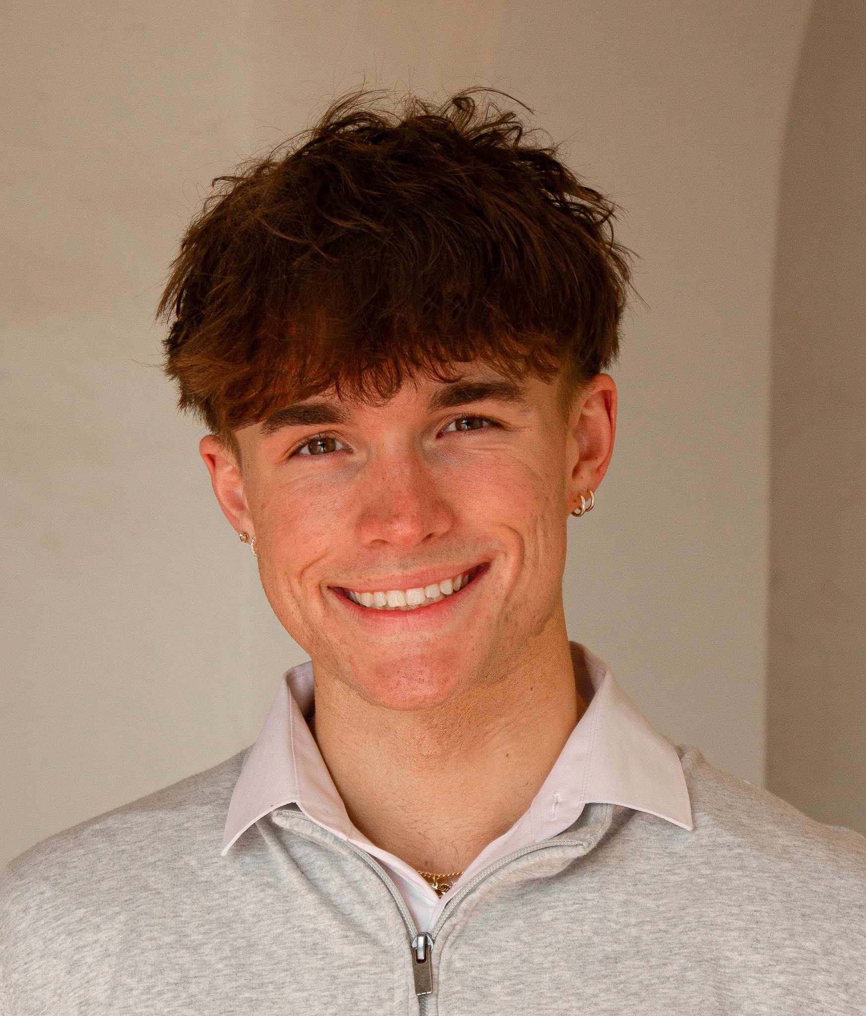 A young man with short, wavy brown hair and earrings, smiling at the camera. He is wearing a light gray zip-up sweater over a white collared shirt, standing indoors with a plain background.