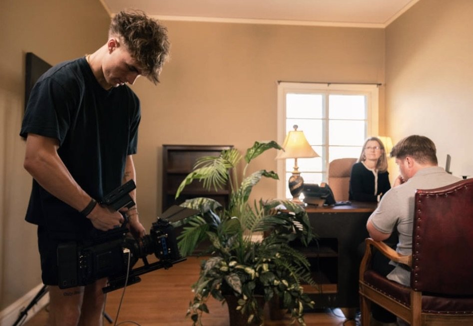 A young man operating a professional camera on a stabilizer while a woman and a man sit at a desk in a home office. The woman is looking at the man, who appears to be distressed and covering his face.