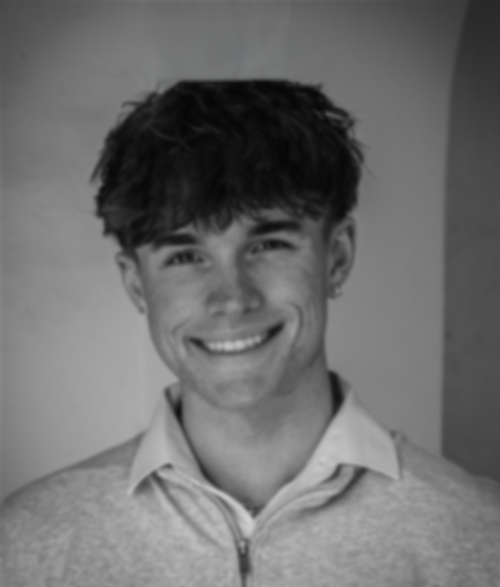 Black and white photo of a young man with wavy hair, smiling, wearing a collared shirt and a zip-up sweater.