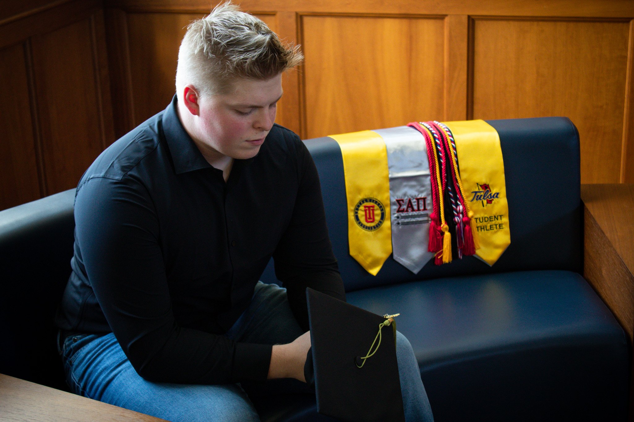 A young man sitting on a black couch, looking down at a graduation cap. Behind him on the couch are yellow and gray honor cords and banners from a university and student organization.