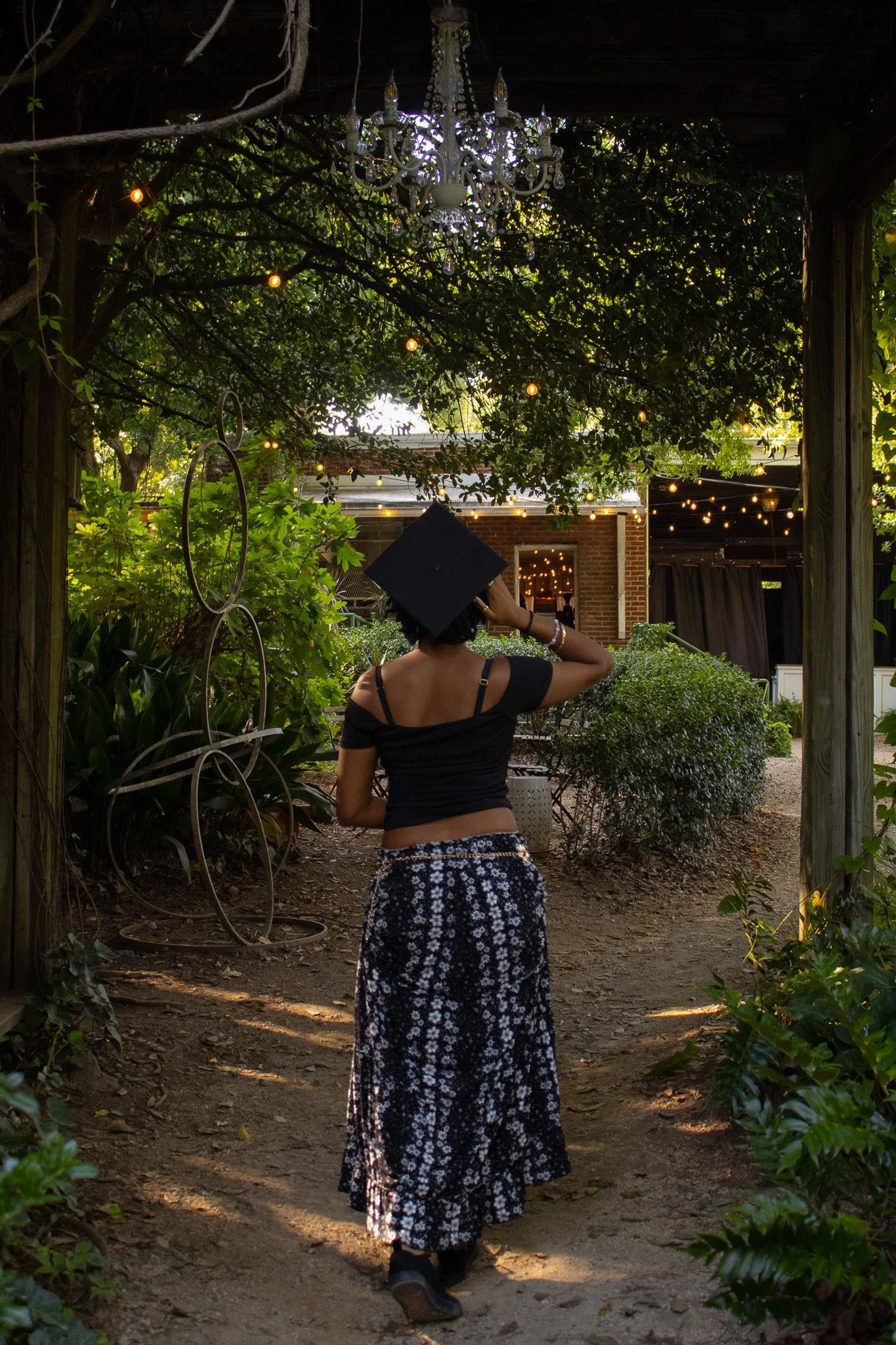 A woman wearing a graduation cap and casual clothes walking through a garden with greenery and string lights.