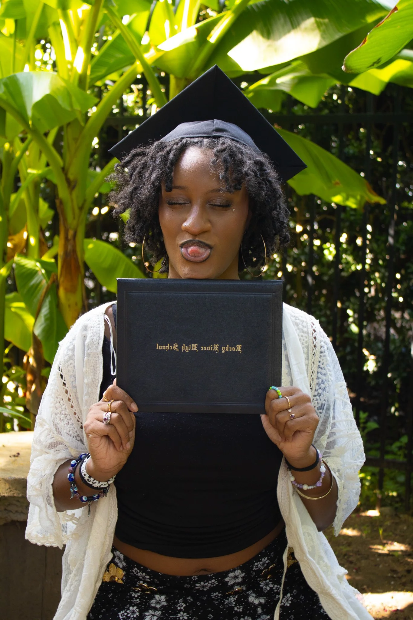 A woman wearing a graduation cap and holding a diploma, making a playful face outdoors with green tropical plants in the background.