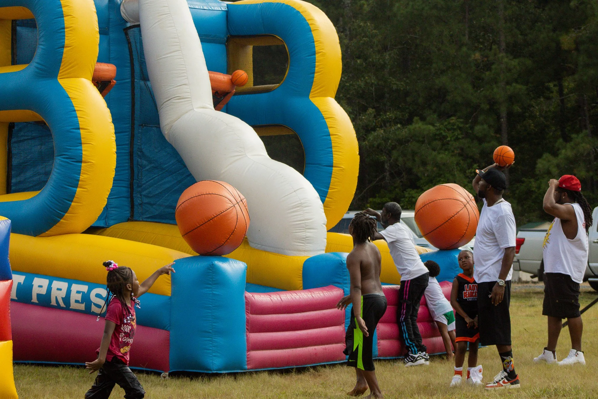 Children playing on an outdoor inflatable bounce house with basketball hoops and giant orange basketballs during the daytime.