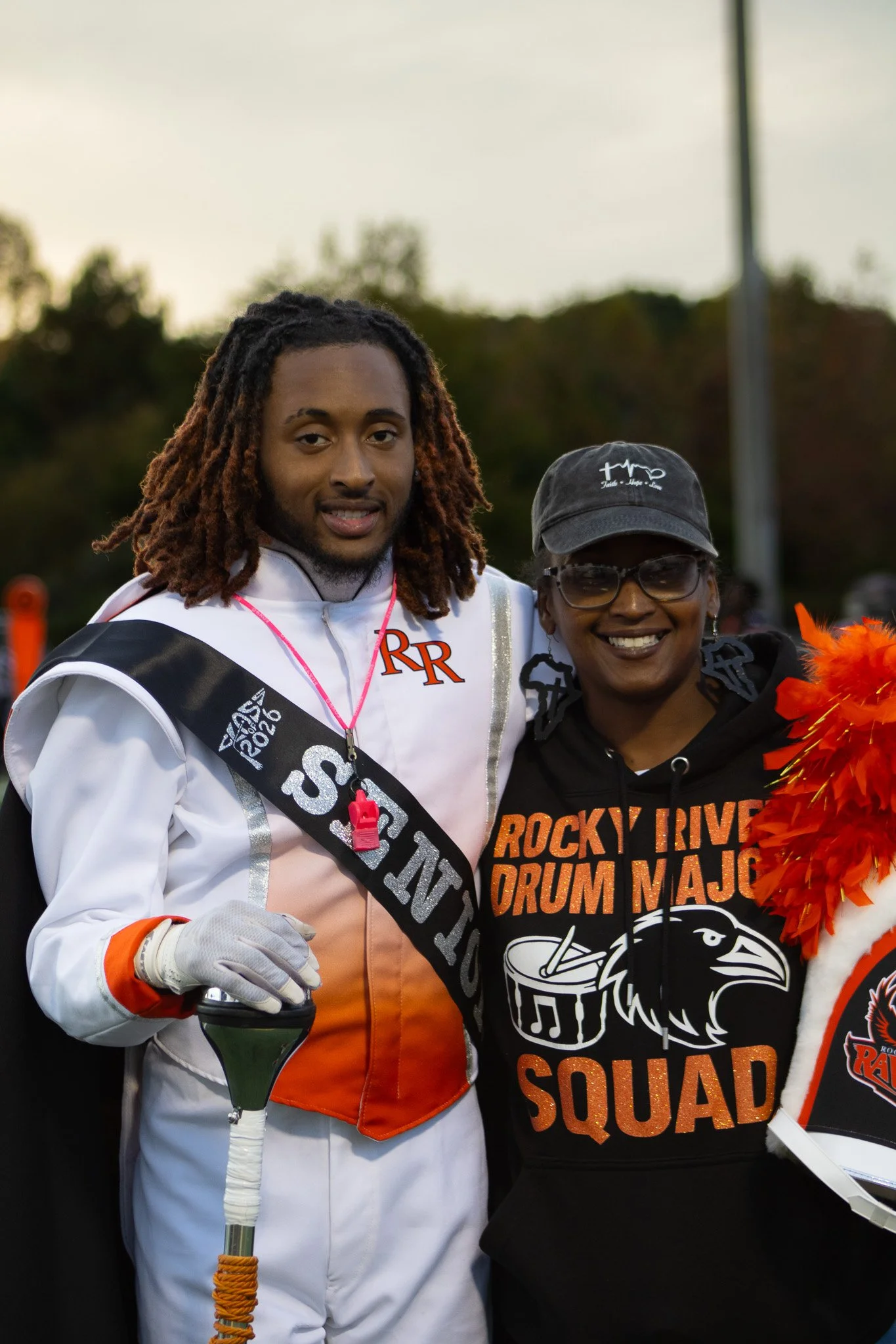 A young man dressed in a white and orange football uniform with a black sash and a woman in a black hoodie with orange and white lettering and a black cap, standing outdoors at sunset, smiling and posing together.