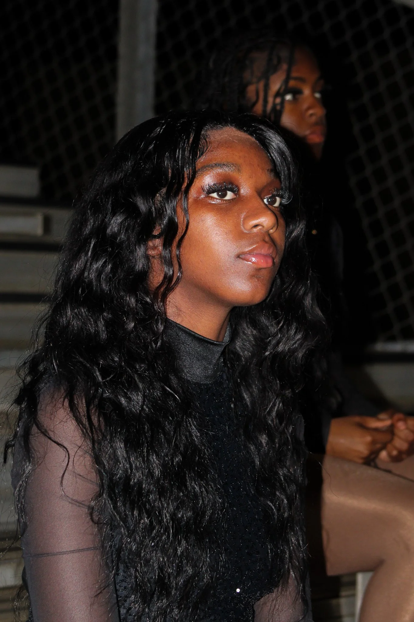 Two women sitting on bleachers at night, one in the foreground with long, curly black hair and shiny makeup, wearing a black sheer top, and one in the background with braided hair, wearing a black outfit.