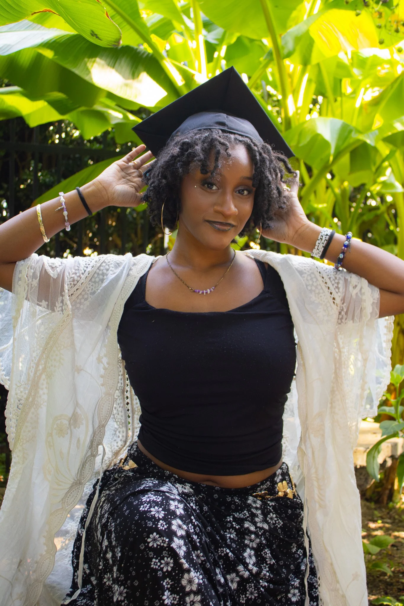 A young woman wearing a black graduation cap, a black top, and patterned pants, standing outdoors with lush green foliage background, smiling and adjusting her cap.
