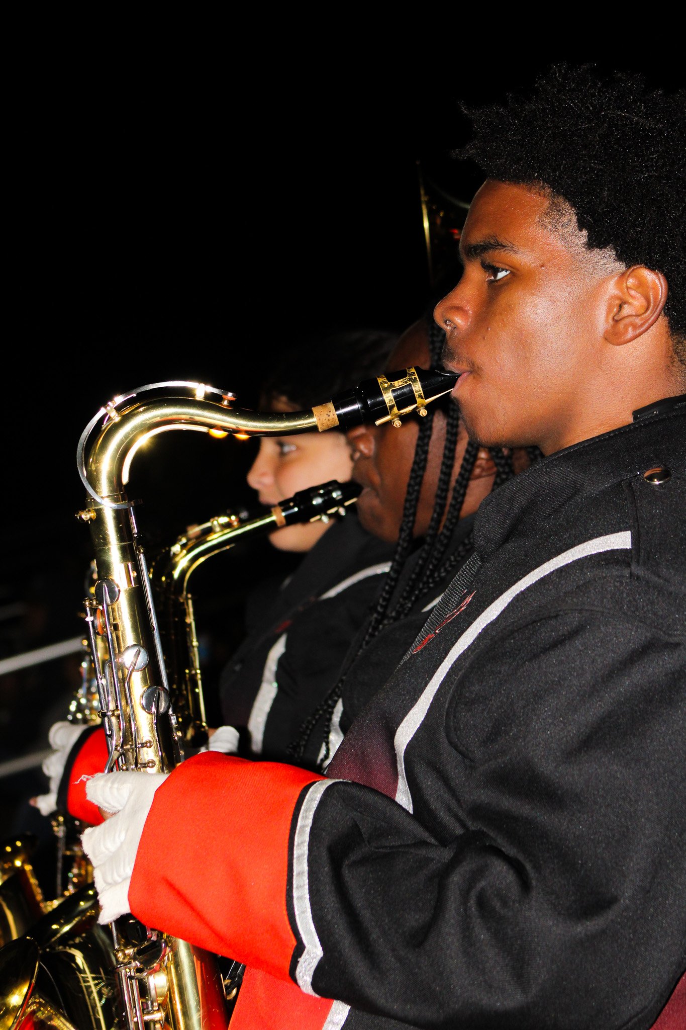Young man playing a saxophone during a performance, with other band members visible in the background.